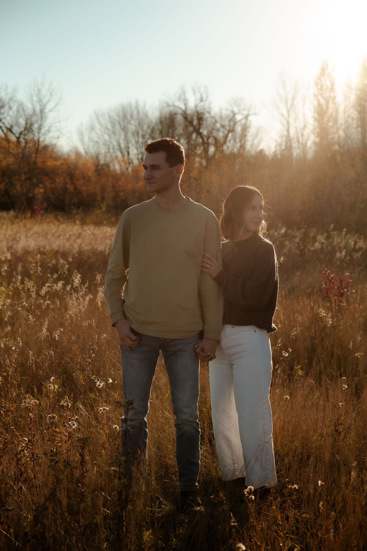 Couple holding hands in golden autumn field at sunset, man in beige sweater and jeans, woman in dark top and white pants.