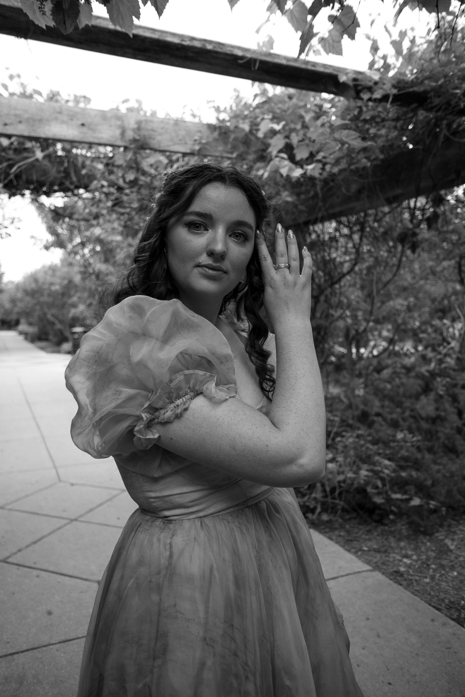 Black and white portrait of a young woman in a ruffled organza dress posing under a garden pergola.