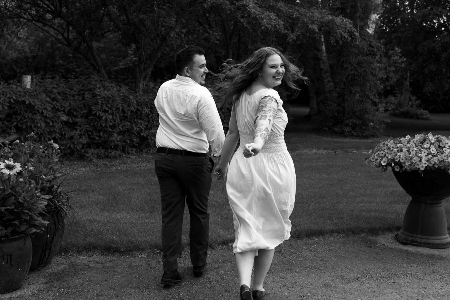 Black and white photo of a couple holding hands and walking through a lush garden, woman in white dress with hair blowing.