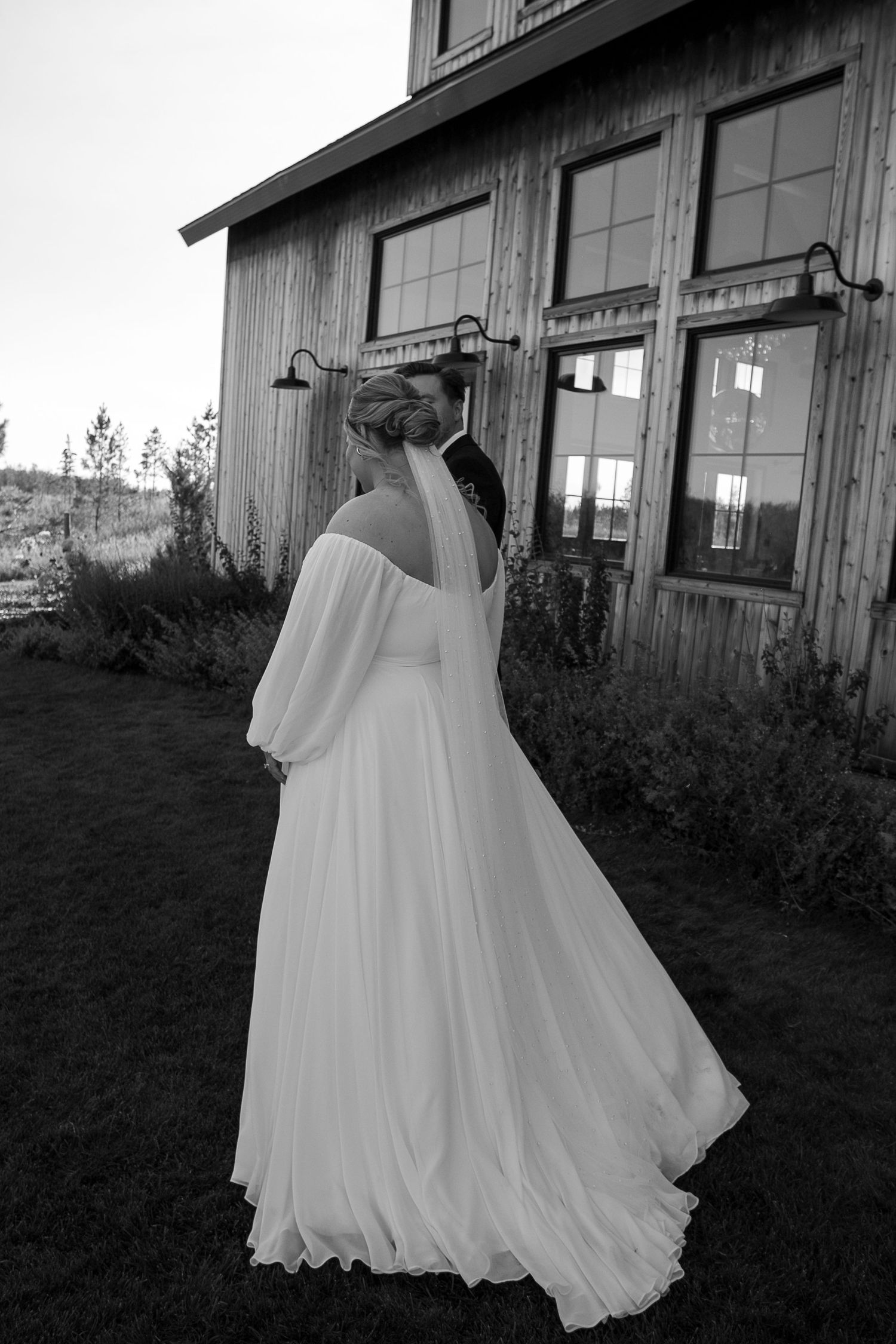 Flowing white wedding dress captured in black and white against rustic building exterior at the Barn at Wind's Edge in Saskatoon, Saskatchew