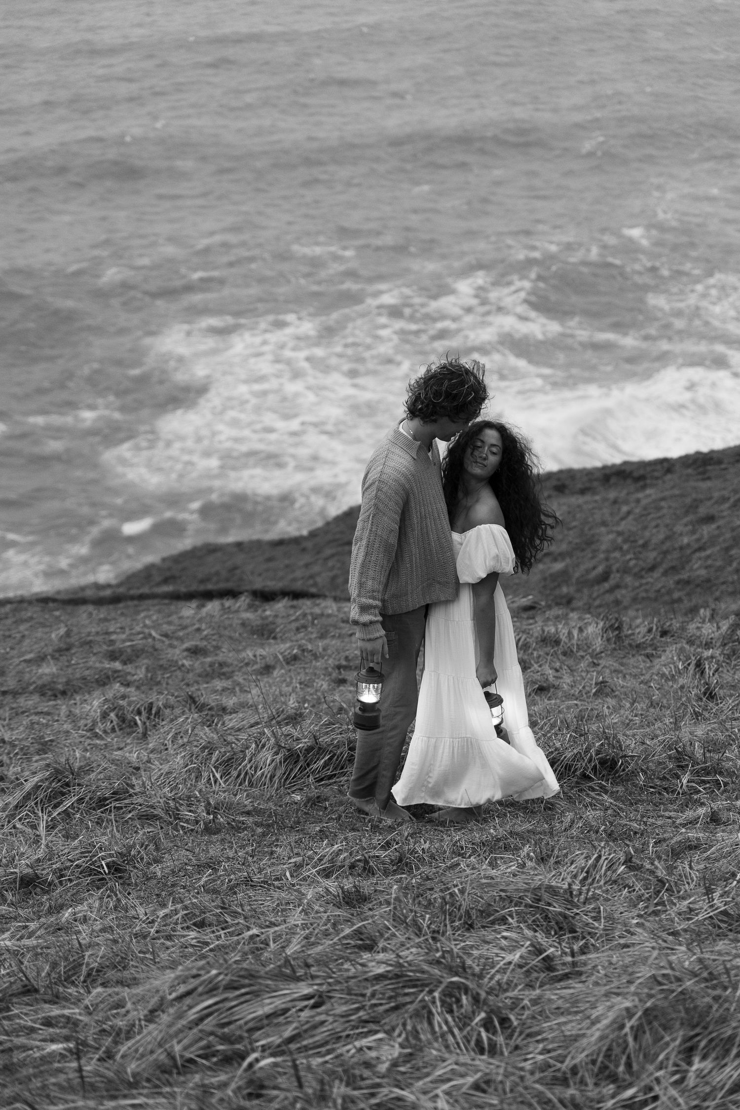 Couple embraces on coastal cliff with ocean waves crashing below.