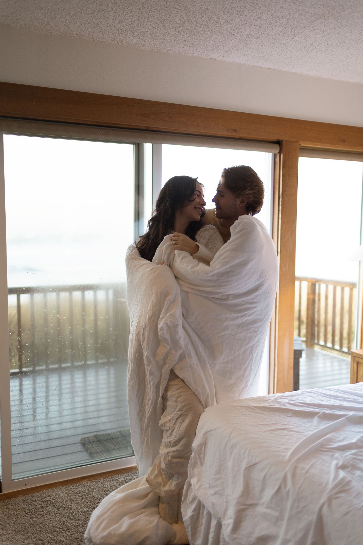 Couple wrapped in white duvet embracing by large bedroom window with ocean view balcony.