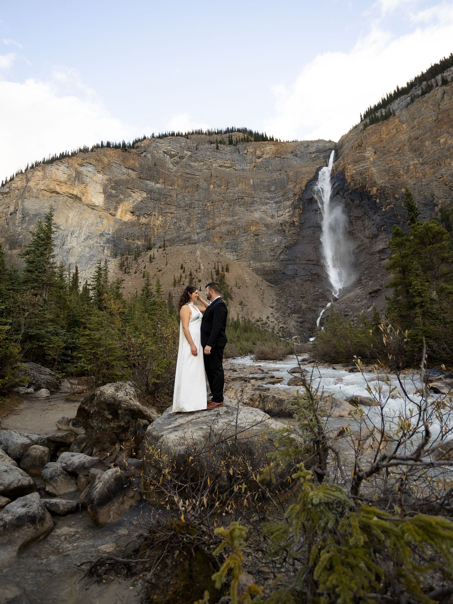 Couple in wedding attire kissing on rocks before Takakkaw Falls waterfall and mountain backdrop in Yoho National Park.
