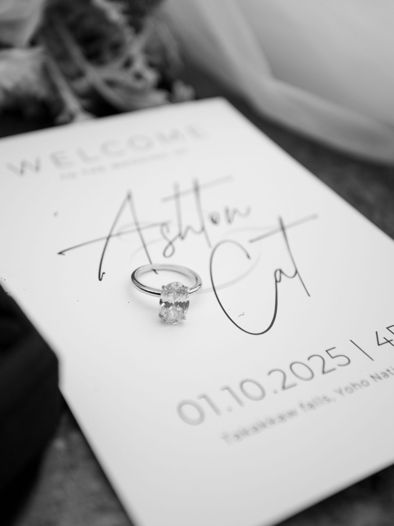 Black and white photo of a diamond engagement ring resting on a wedding welcome sign dated 01.10.2025.