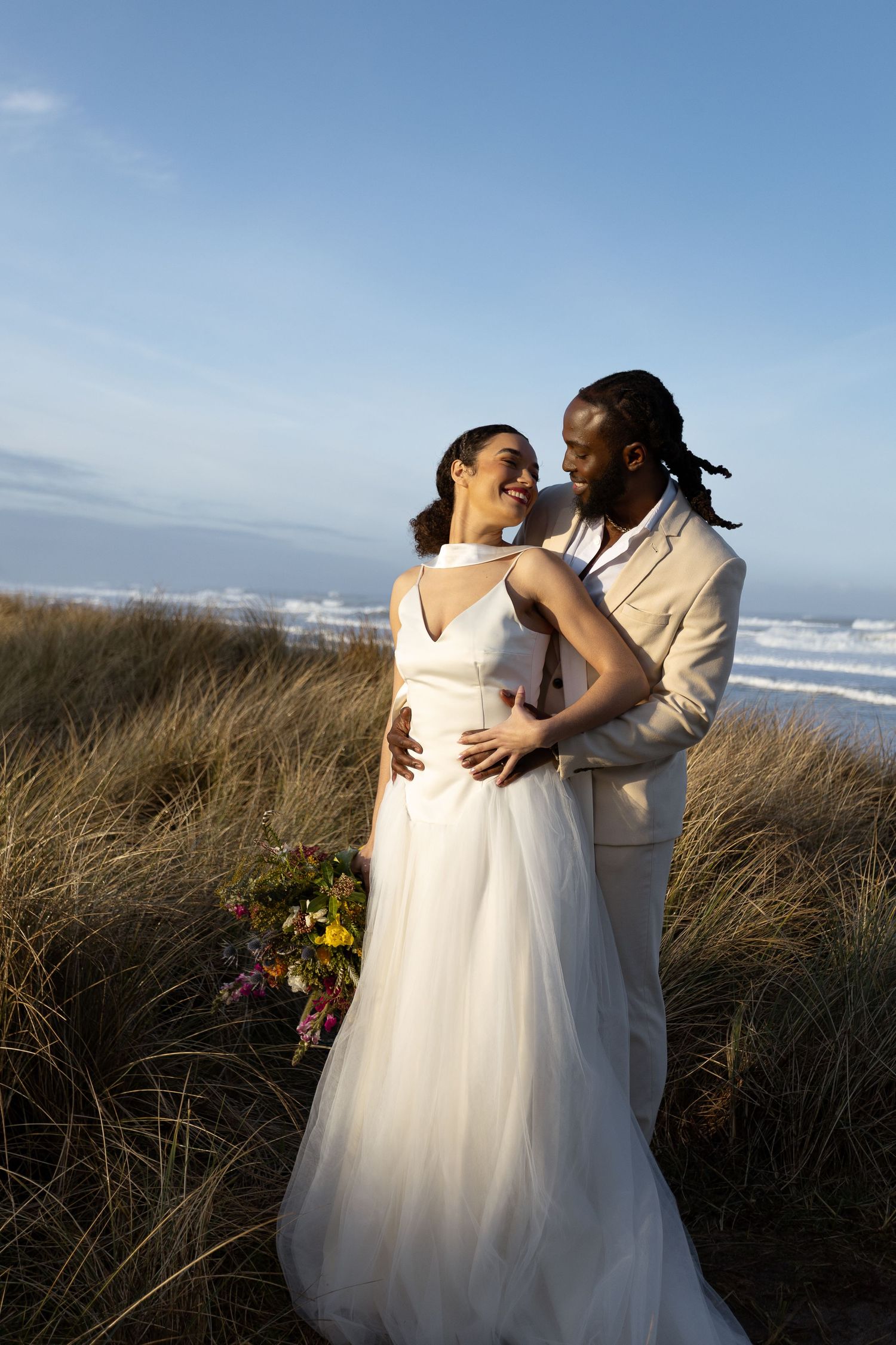 Couple in wedding attire embracing on a beach, bride in white gown and groom in tan suit with dreadlocks.