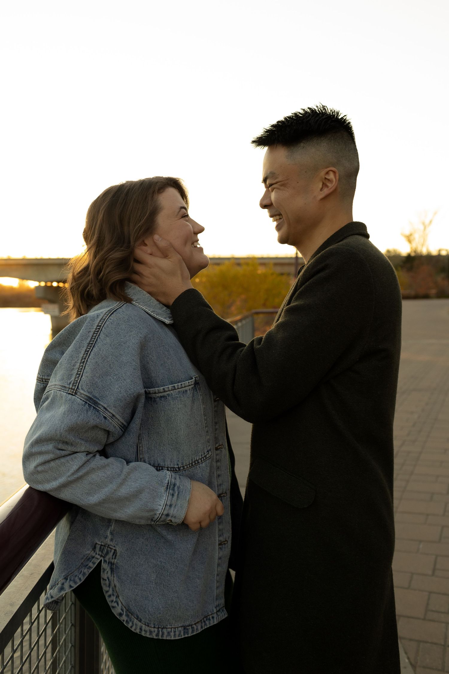 A smiling couple sharing a romantic moment by a riverside railing at sunset, woman in denim jacket, man in dark coat.
