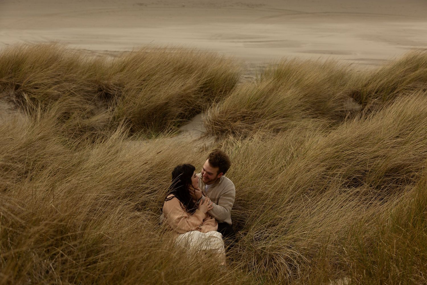 Couple embracing romantically among tall golden beach grass dunes in a moody, intimate outdoor engagement photo.
