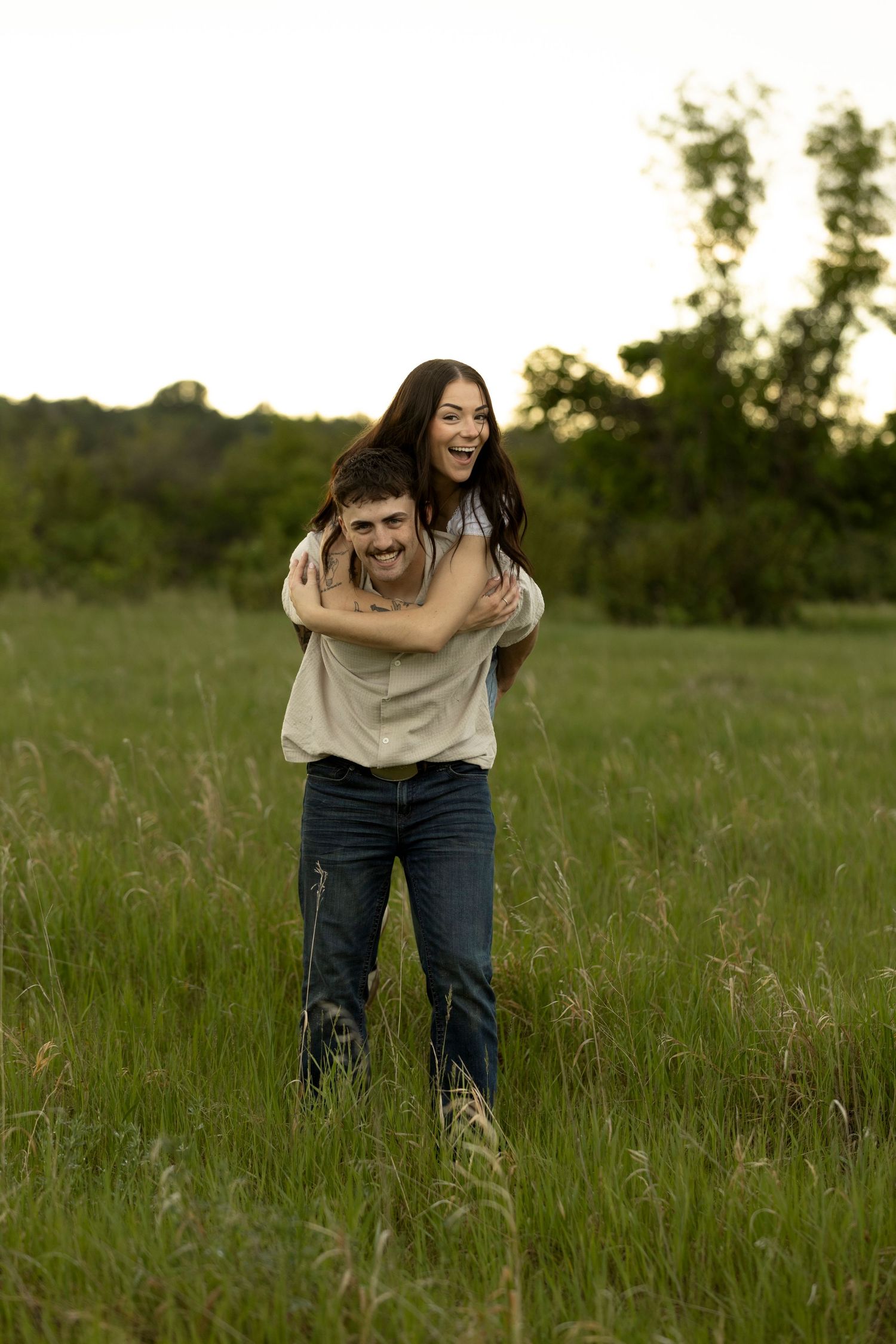 A joyful couple laughing in a green field, man giving woman a piggyback ride at sunset.