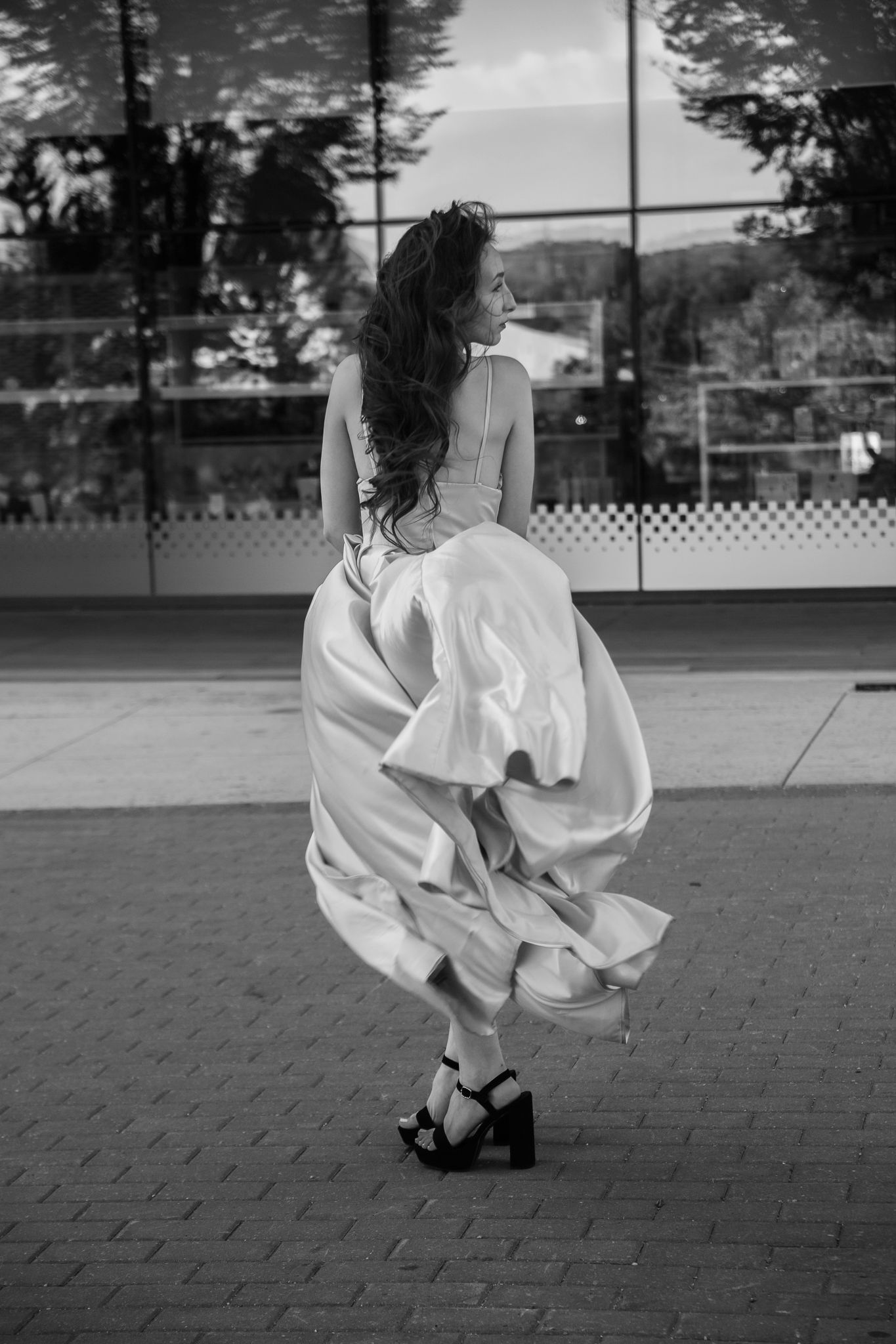 Black and white photo of a woman in a flowing white gown and heels, twirling outdoors near a glass building.