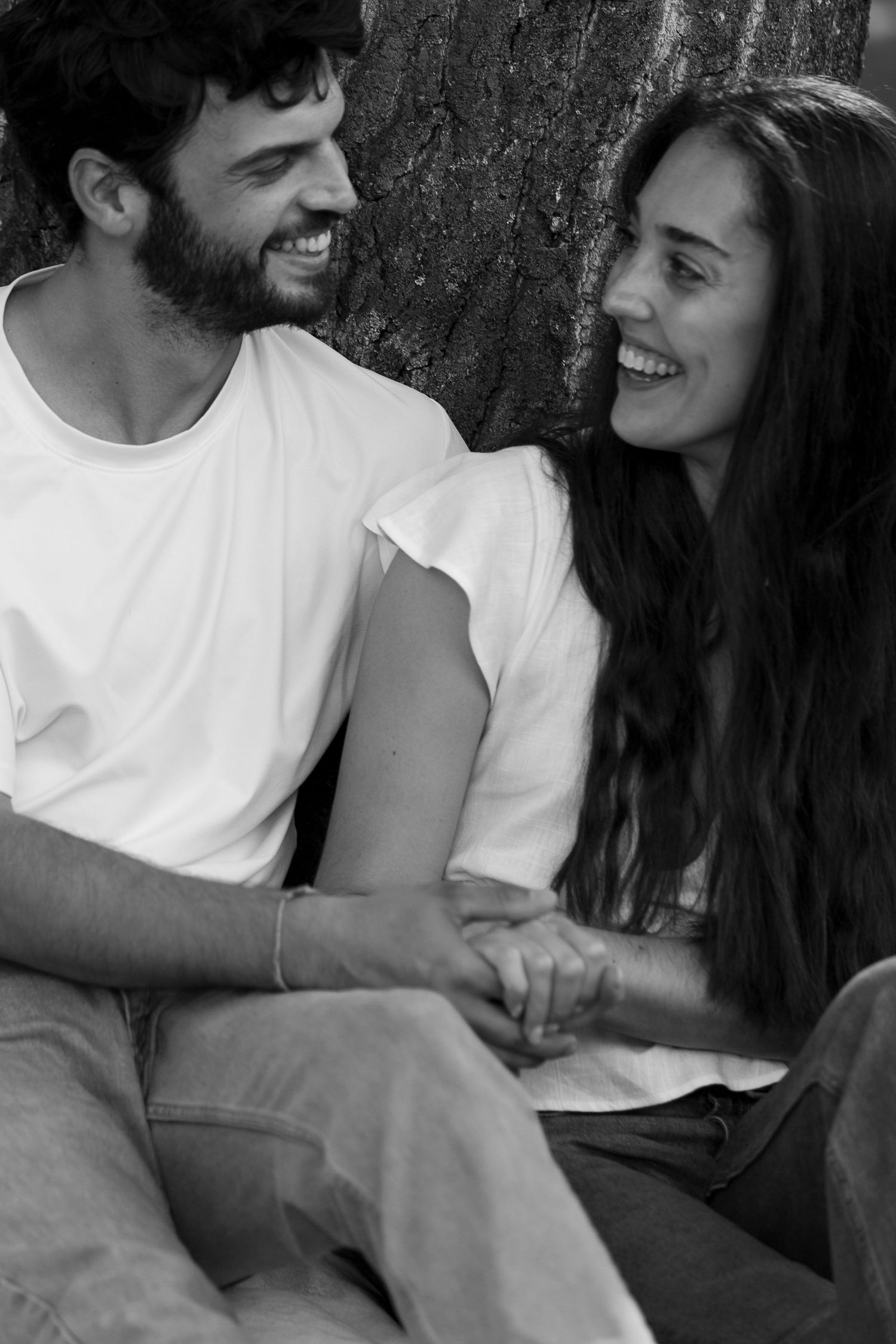 Black and white photo of a smiling couple holding hands while sitting together against a tree.
