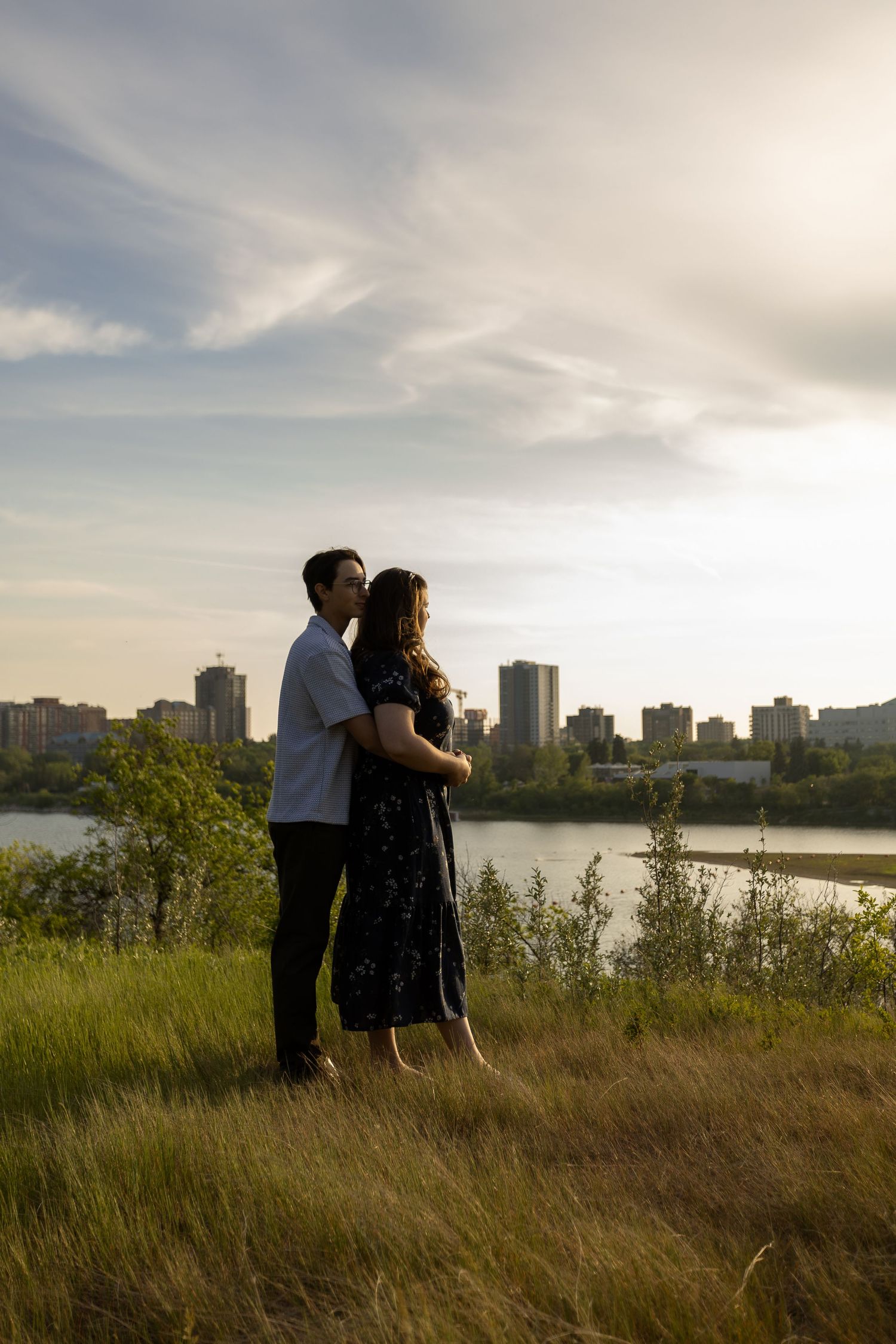 Couple embracing on a grassy hill with a city skyline and river in the background at sunset.