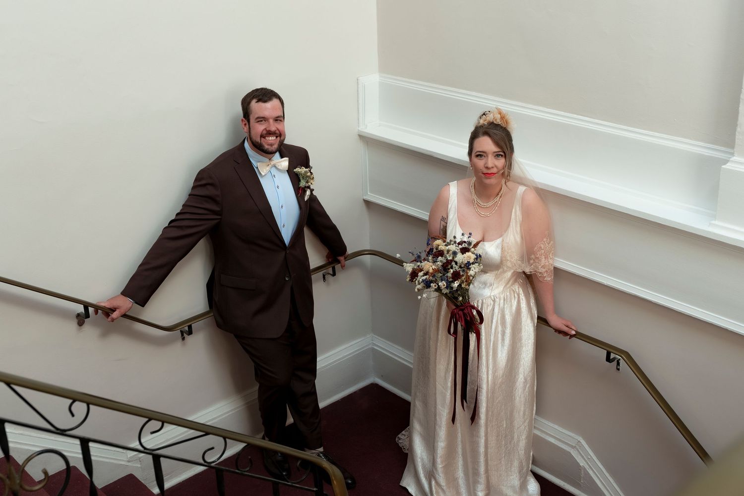 Bride and groom posing on elegant staircase, bride holding bouquet in vintage gown, groom in brown suit with boutonniere.