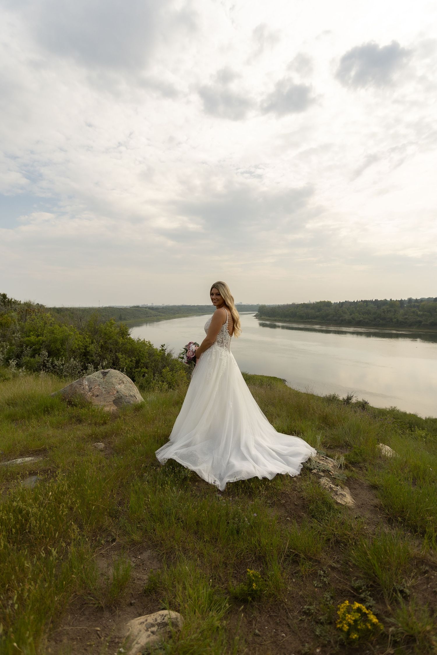 A flowing white wedding dress twirls against a scenic riverside landscape with green hills and cloudy skies.