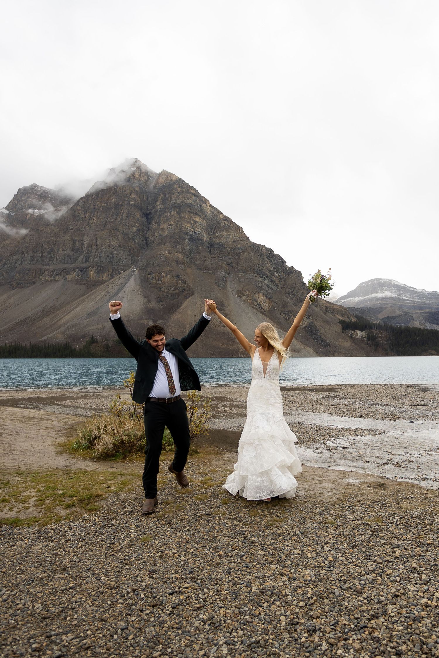 Joyful newlyweds celebrate by a mountain lake, bride in white gown holding bouquet, groom in suit, arms raised in triumph.