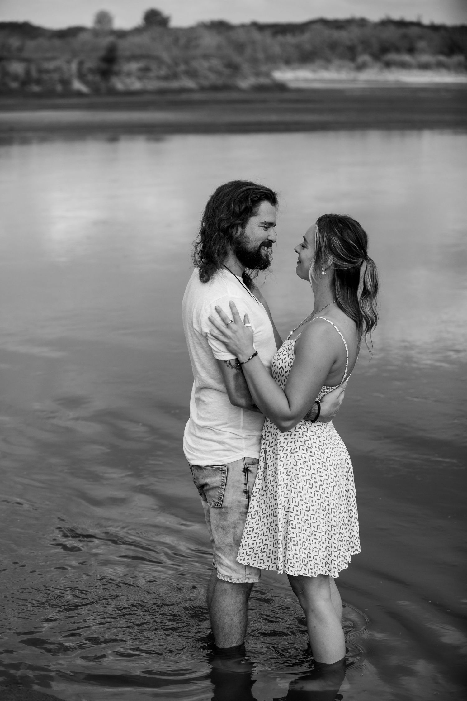 Black and white photo of a romantic couple embracing in shallow water near a lake shore.