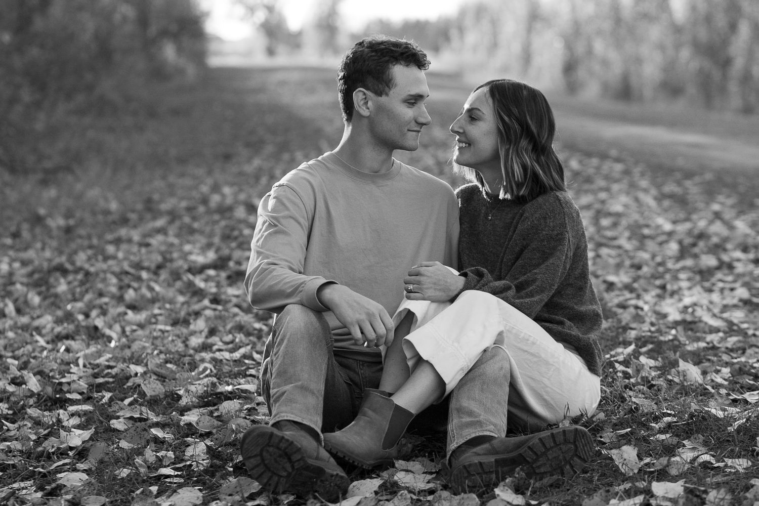 Black and white photo of a romantic couple sitting together on fallen autumn leaves, smiling at each other outdoors.