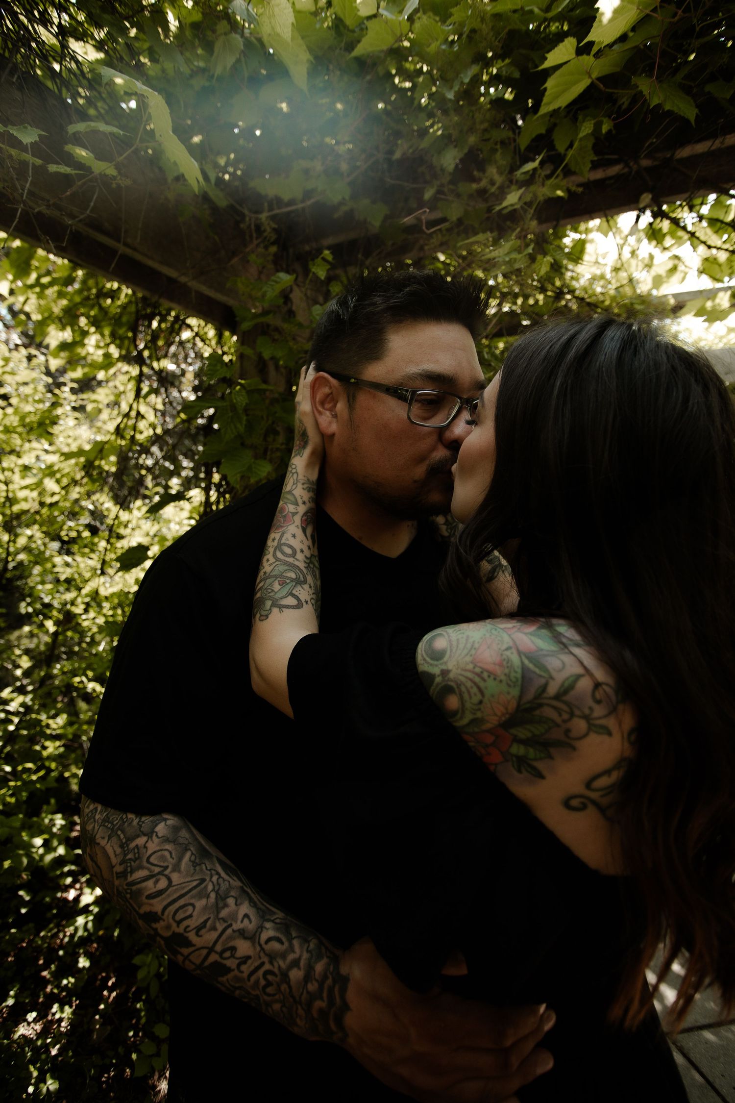 Tattooed couple sharing a kiss under a lush green garden arbor, both wearing black clothing.