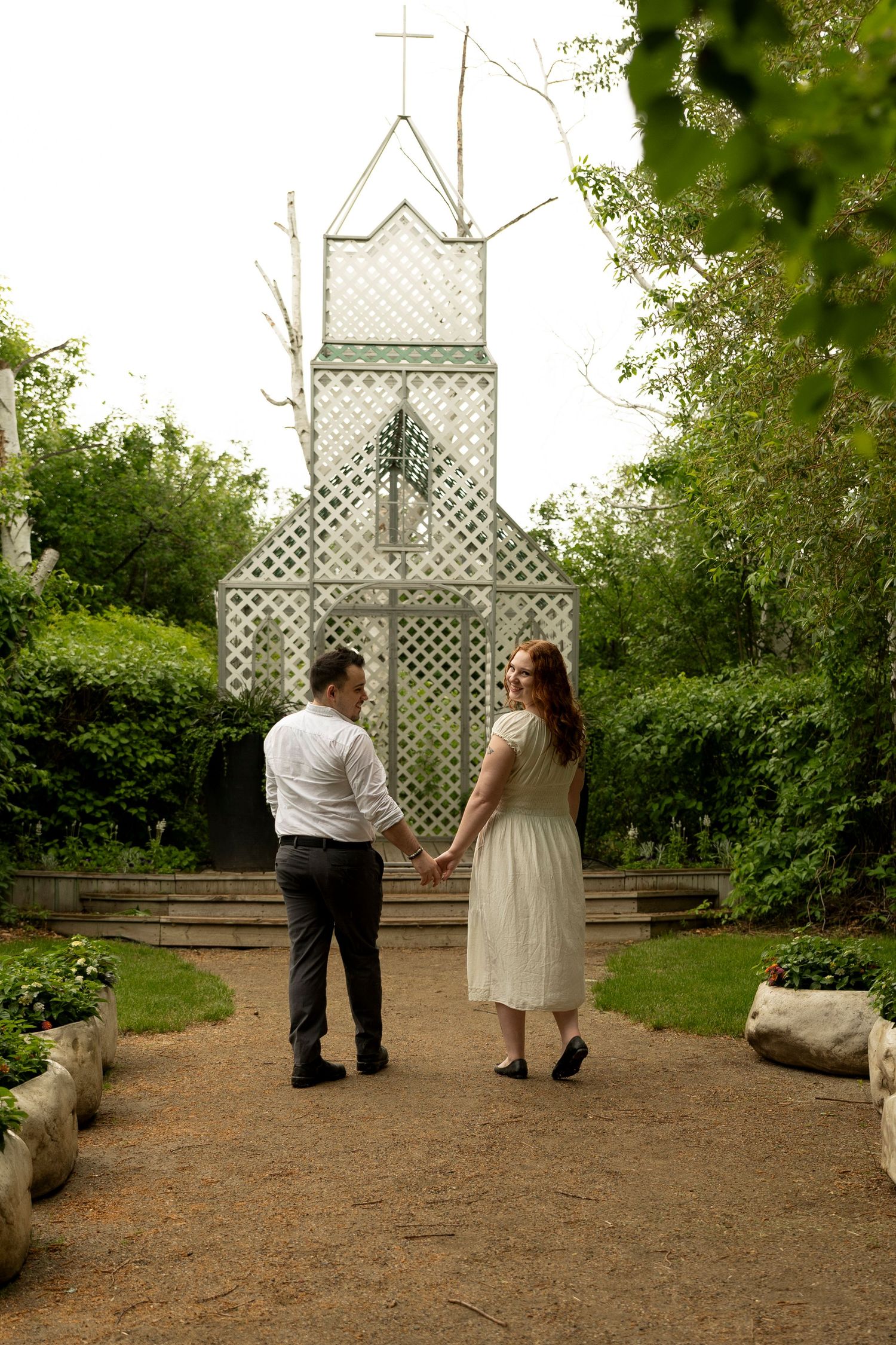 Couple holding hands on garden path before a white lattice chapel surrounded by lush greenery.