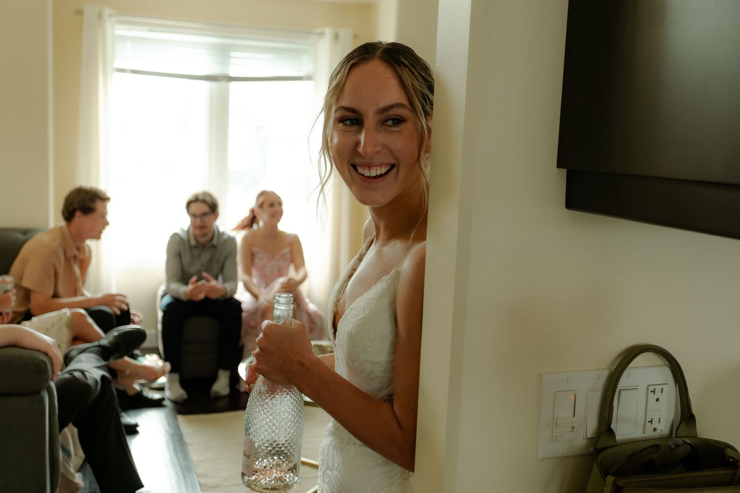 Smiling bride in white dress holding a water bottle, with wedding party guests seated in background.