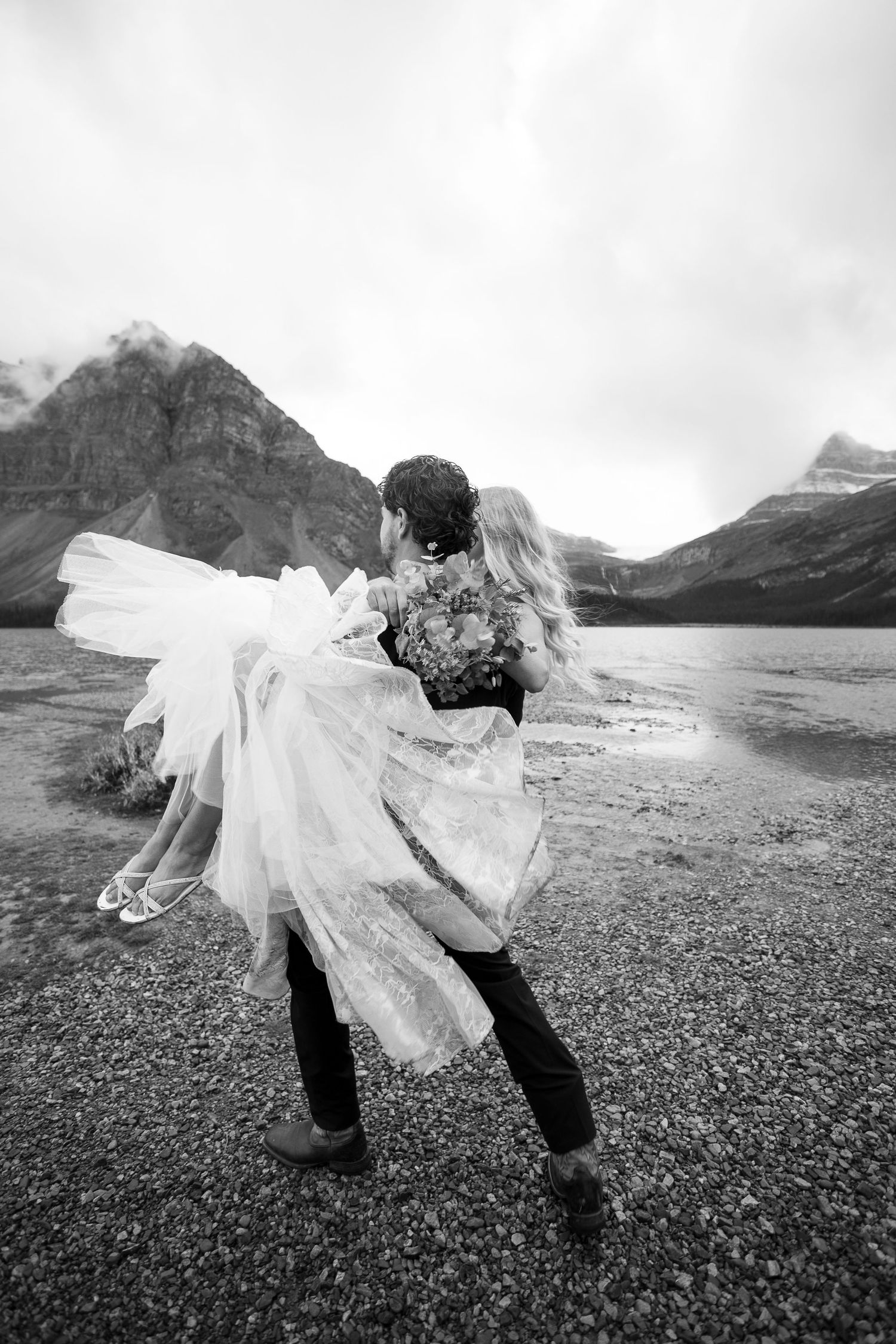 Groom carries bride in wedding dress by a mountain lake in black and white photo.