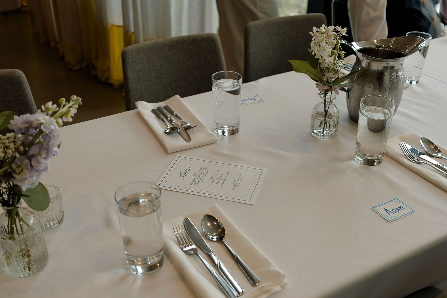 Elegant white tablecloth wedding reception table set with silverware, glassware, floral centerpieces, and folded napkins.