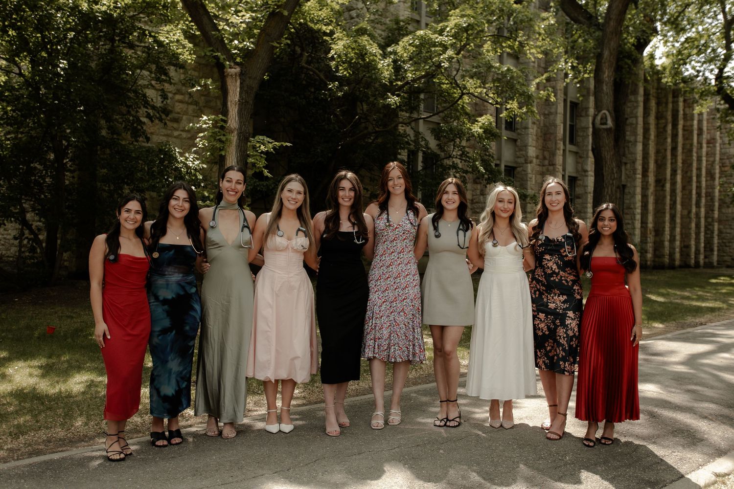 Group of ten women in elegant dresses posing together outdoors under shaded trees for a formal occasion.