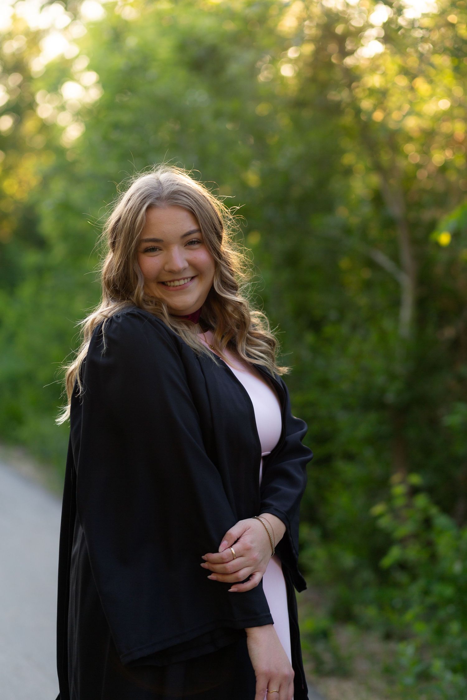 Smiling young woman in black graduation gown over pink dress poses on a tree-lined path in golden hour light.