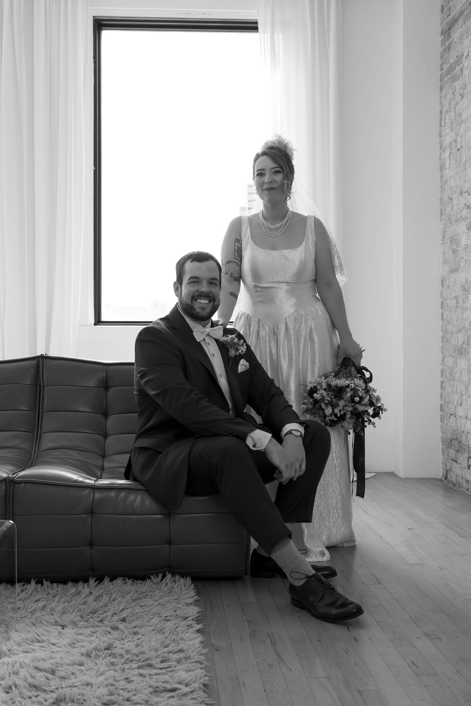 Black and white wedding photo of smiling groom seated on leather sofa with bride standing beside him holding bouquet.