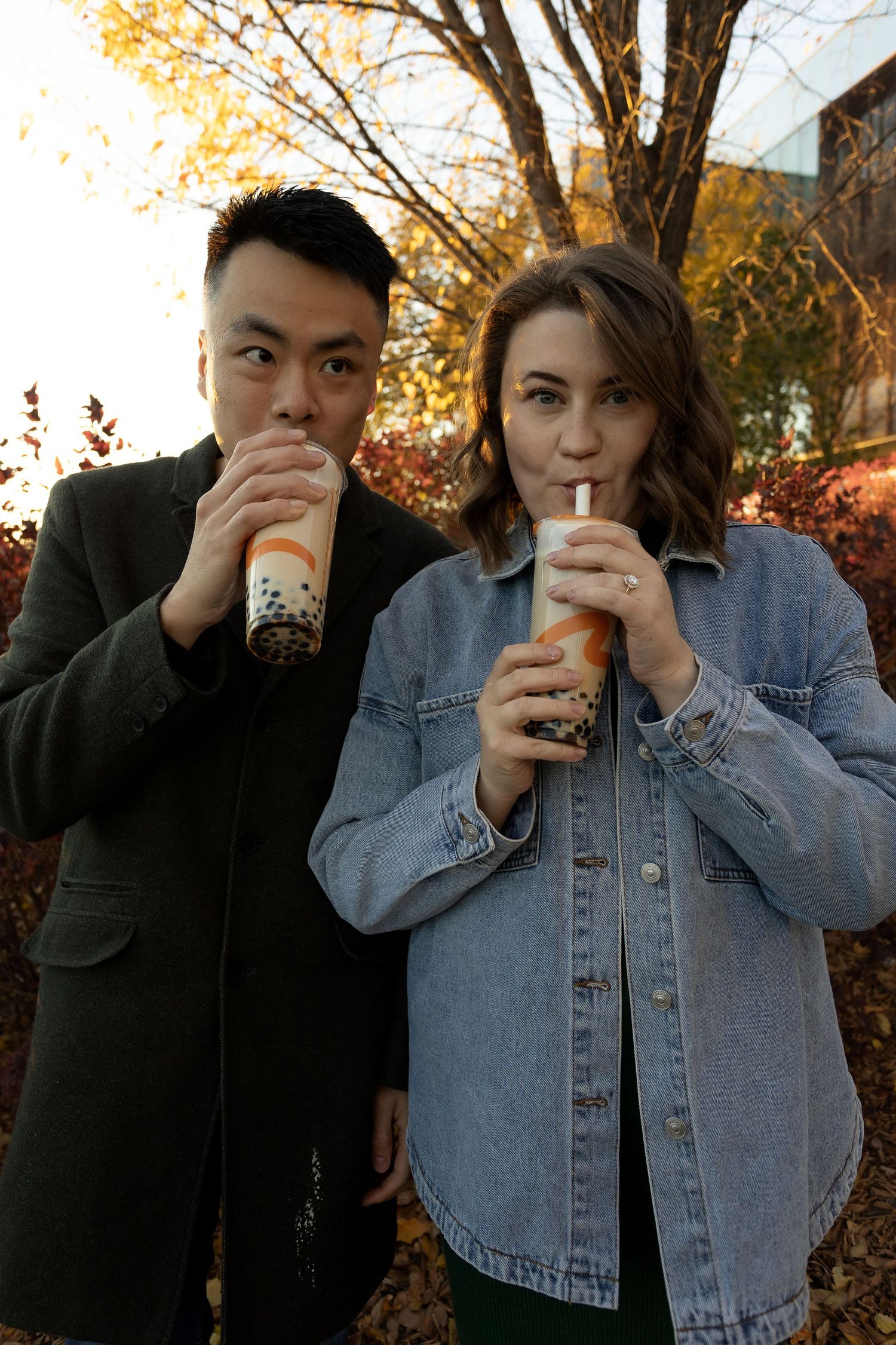Two friends enjoying bubble tea drinks outdoors in autumn, man in green jacket and woman in denim jacket.