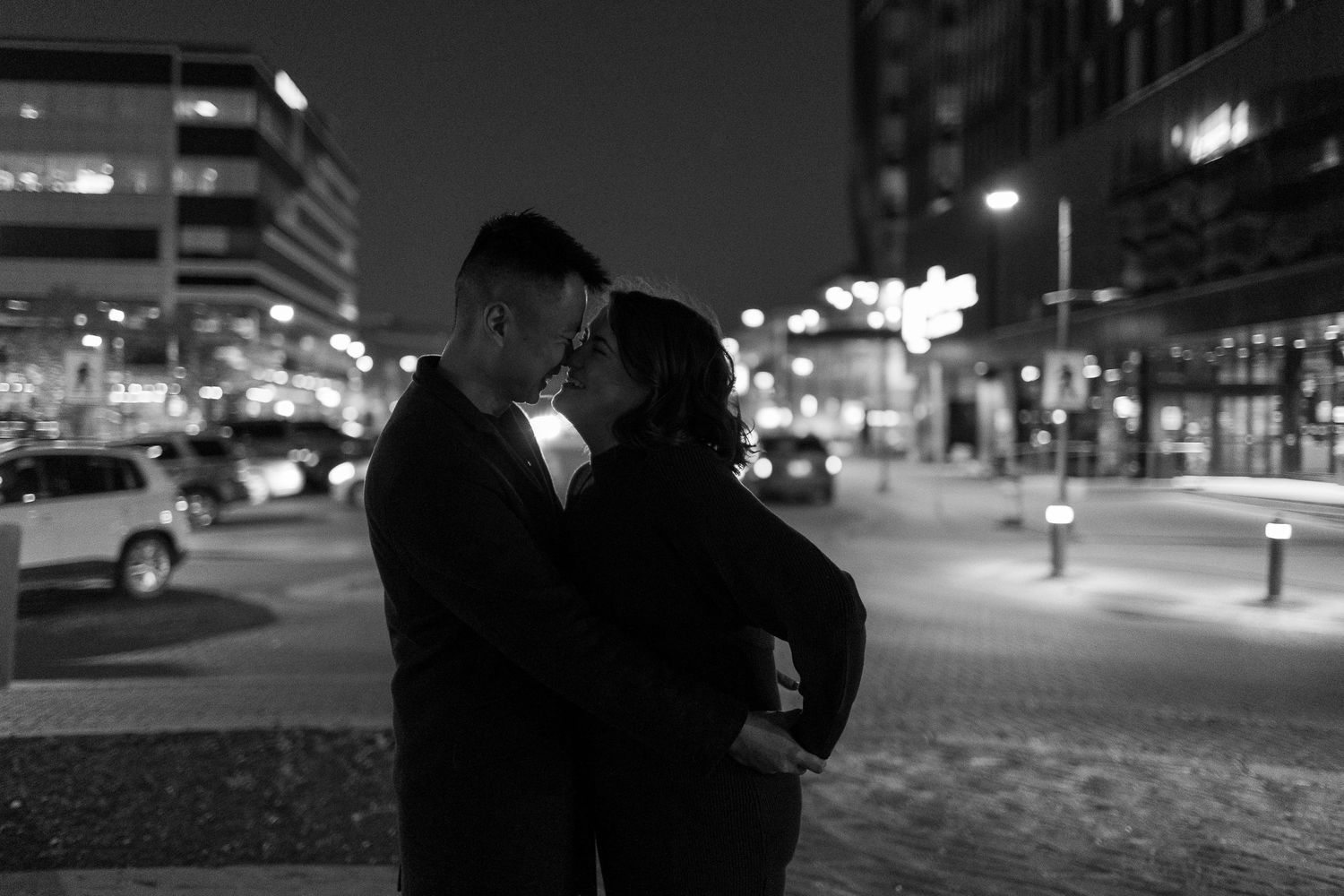 Black and white photo of a couple sharing a romantic kiss on a city street at night with glowing lights behind them.