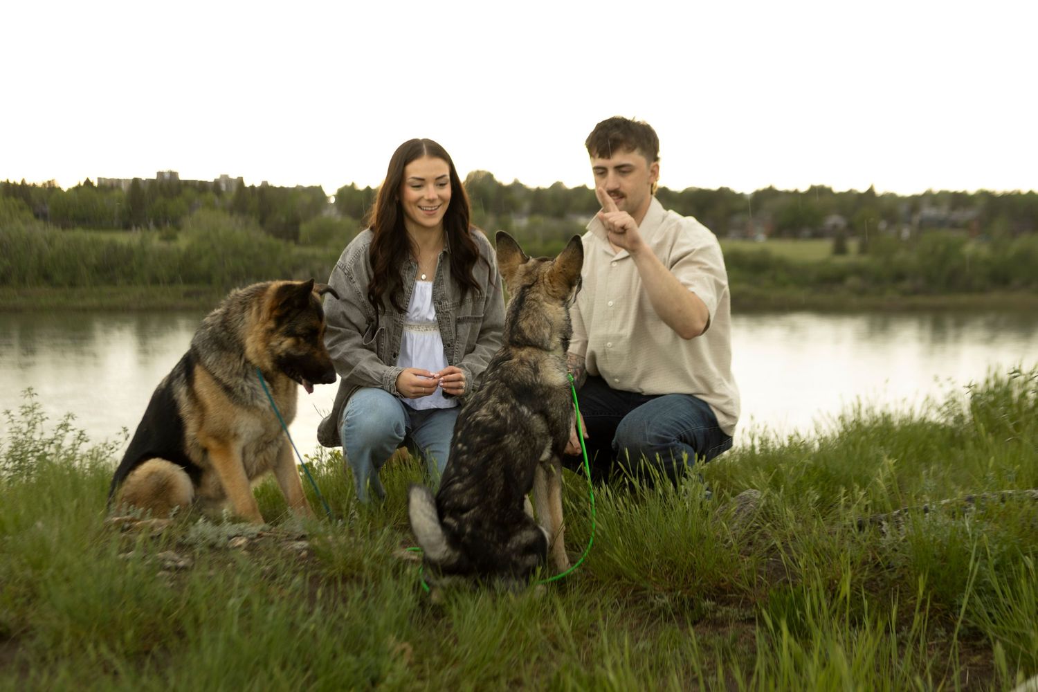 Young couple with two German Shepherds by a lake in a green field, man gesturing while woman smiles warmly.