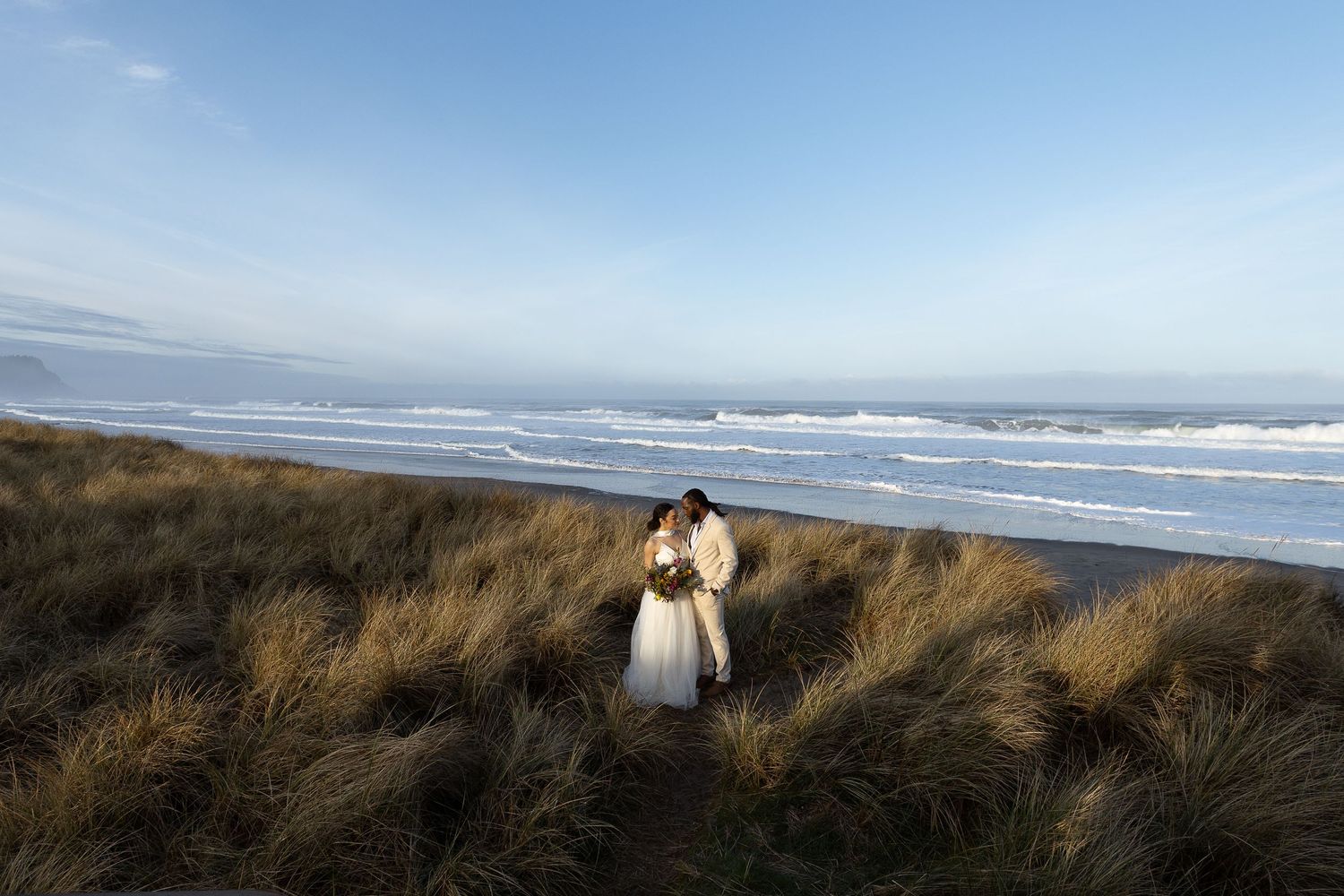 Couple embracing in coastal dune grass with ocean waves in background during a romantic beach wedding.