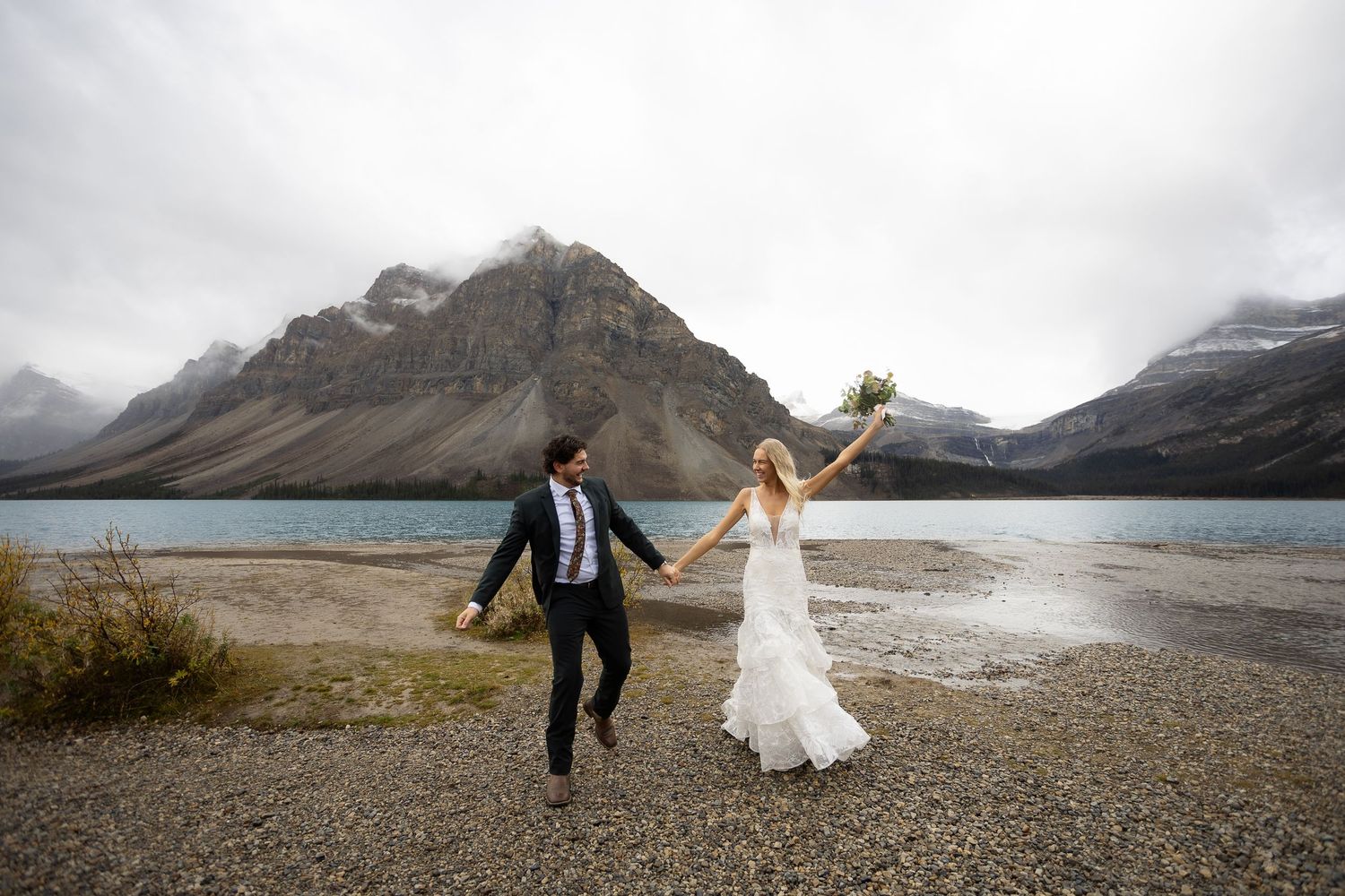Bride and groom hold hands by a mountain lake, bride raises bouquet joyfully, stunning Rocky Mountain wedding backdrop.