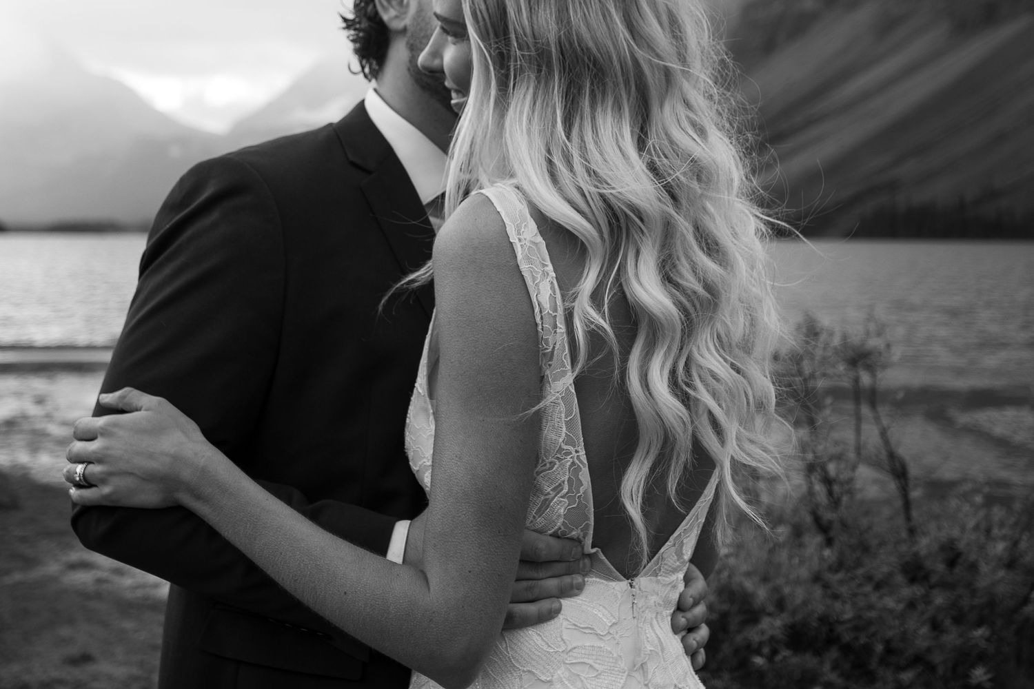 Black and white photo of a bride and groom embracing by a mountain lake, showcasing lace wedding dress back.