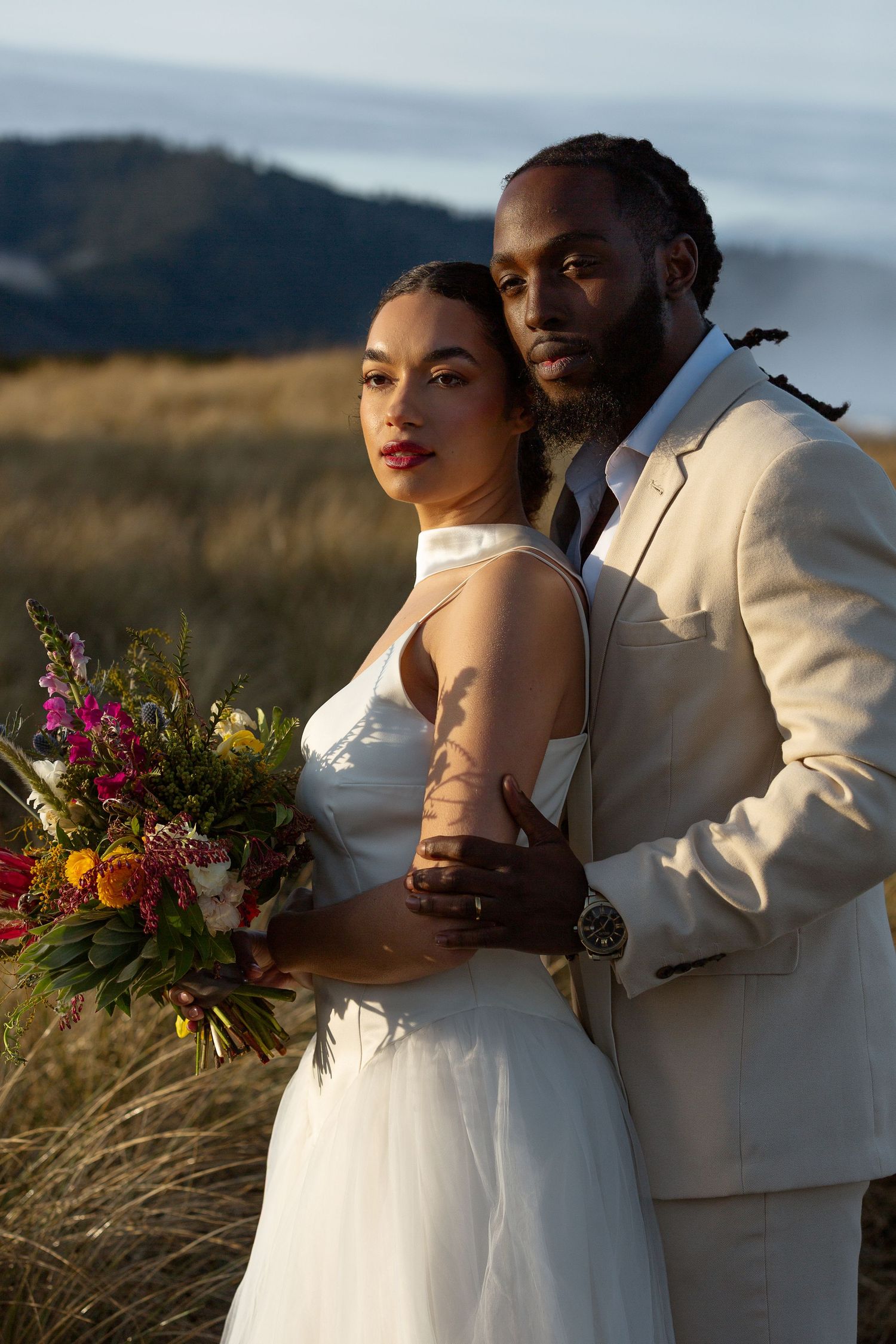 Interracial couple in wedding attire pose outdoors, bride in white gown holding colorful bouquet, groom in beige suit.