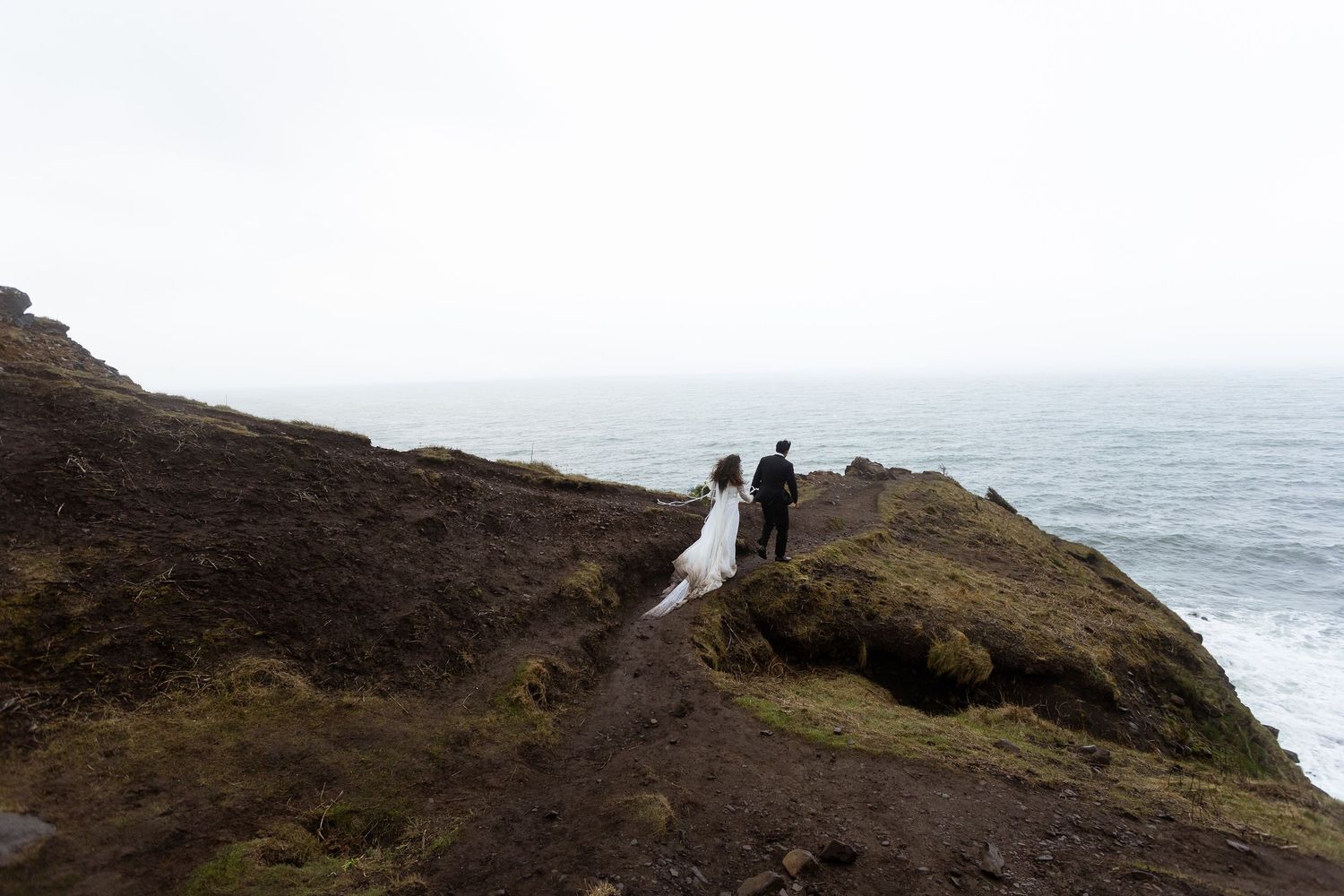 Bride and groom stand on a rugged coastal cliff in Iceland, overlooking the dramatic misty ocean on their elopement day.