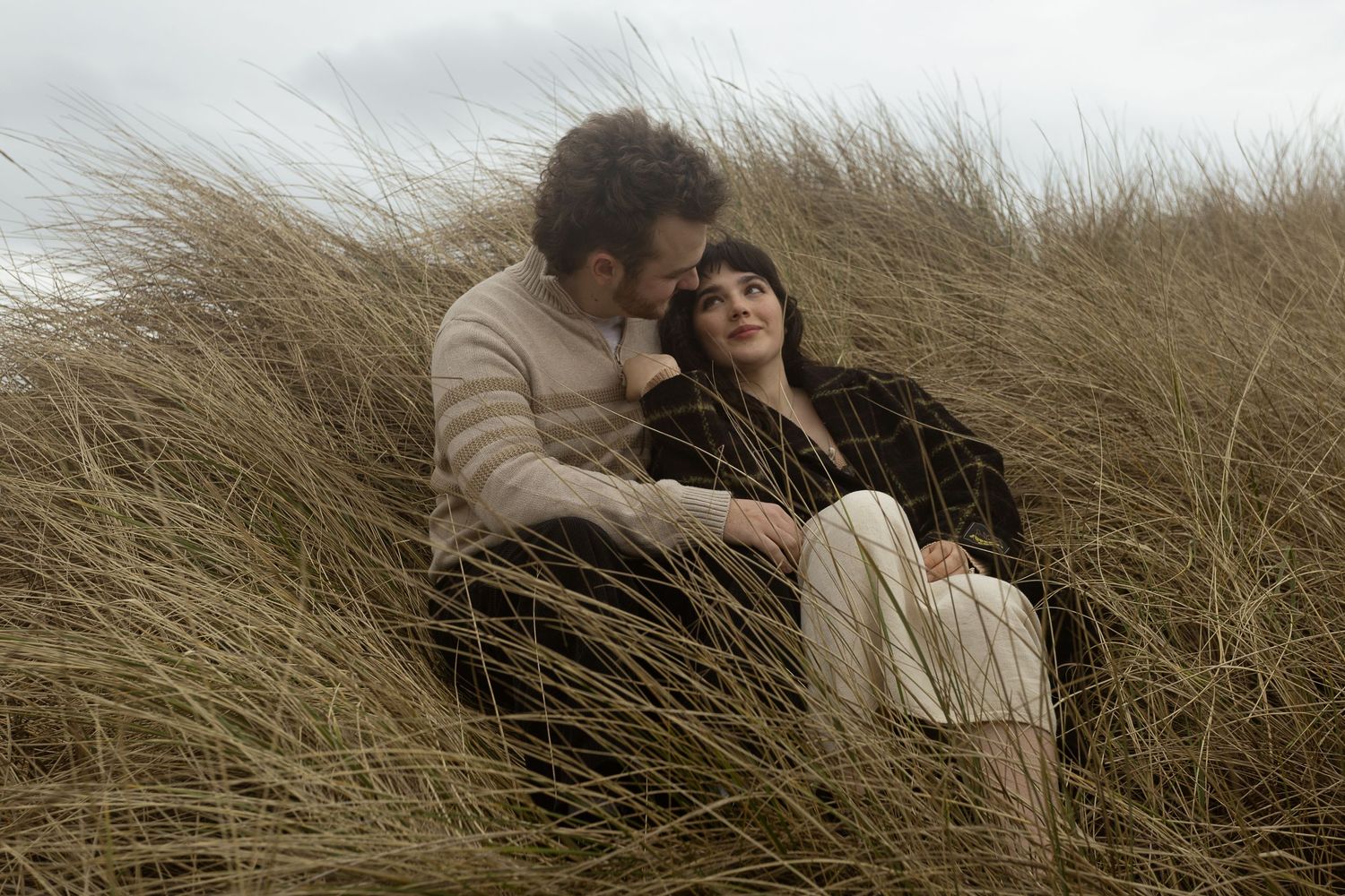 A romantic couple sitting together in tall golden grass dunes, sharing an intimate moment outdoors.