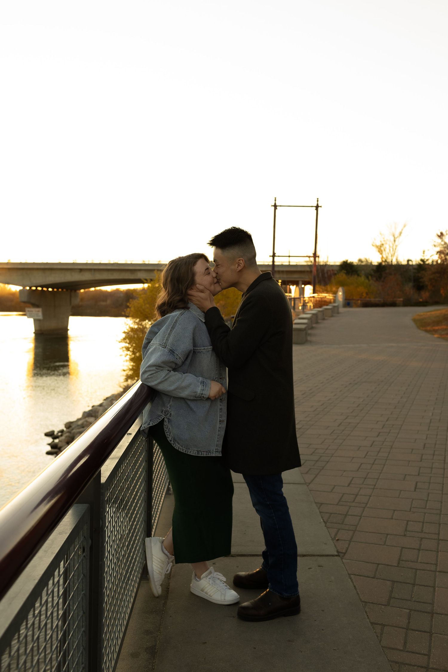A couple shares a romantic kiss on a scenic riverside promenade during a warm golden sunset.