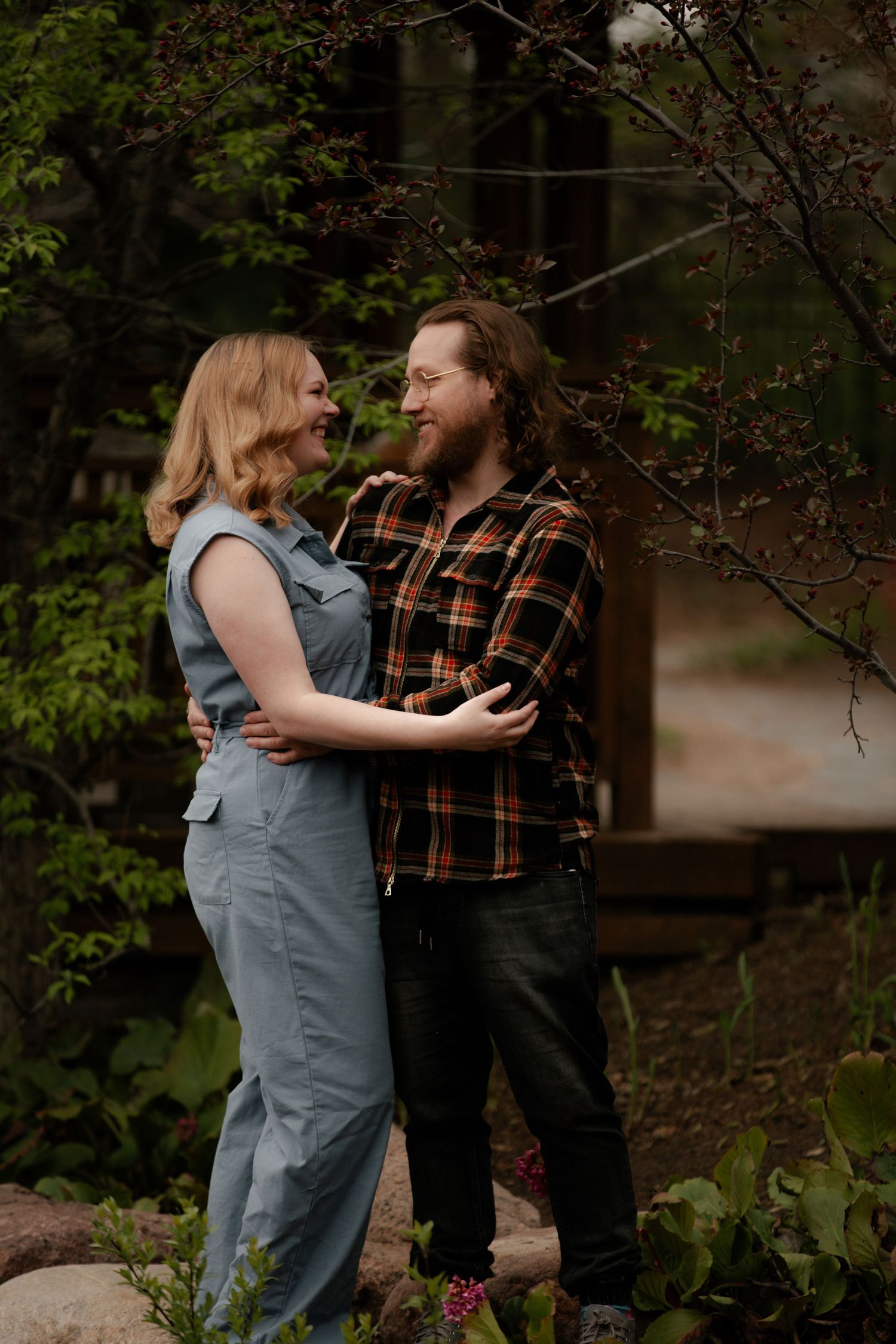 A couple embraces and laughs together outdoors in a lush green forest setting, wearing casual outfits.