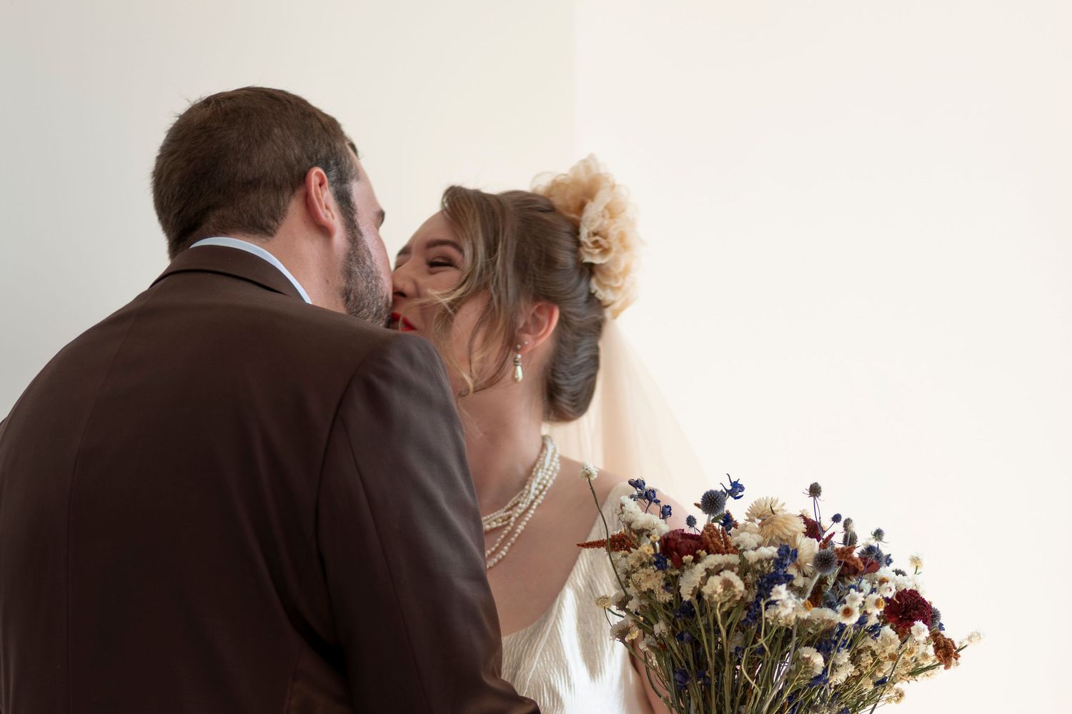 A bride and groom share a romantic kiss, with the bride holding a beautiful bouquet of dried wildflowers in red, blue, and white.