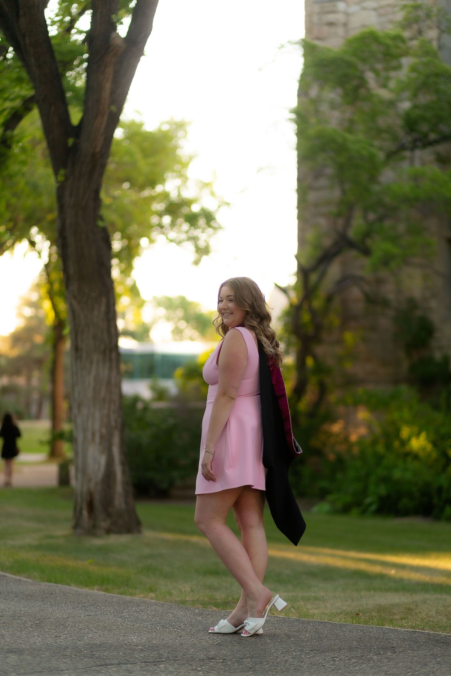 Young woman in pink dress carries black graduation gown while walking on a sunlit campus path near stone building.