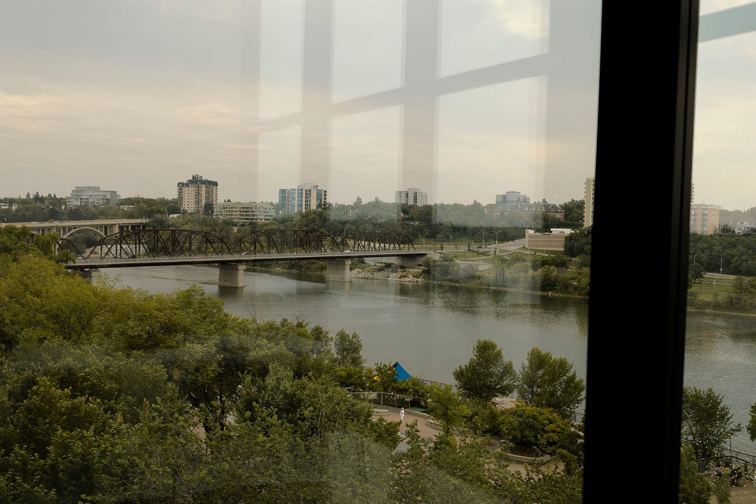 View through glass window of a river bridge and city skyline with lush green trees on a cloudy day.