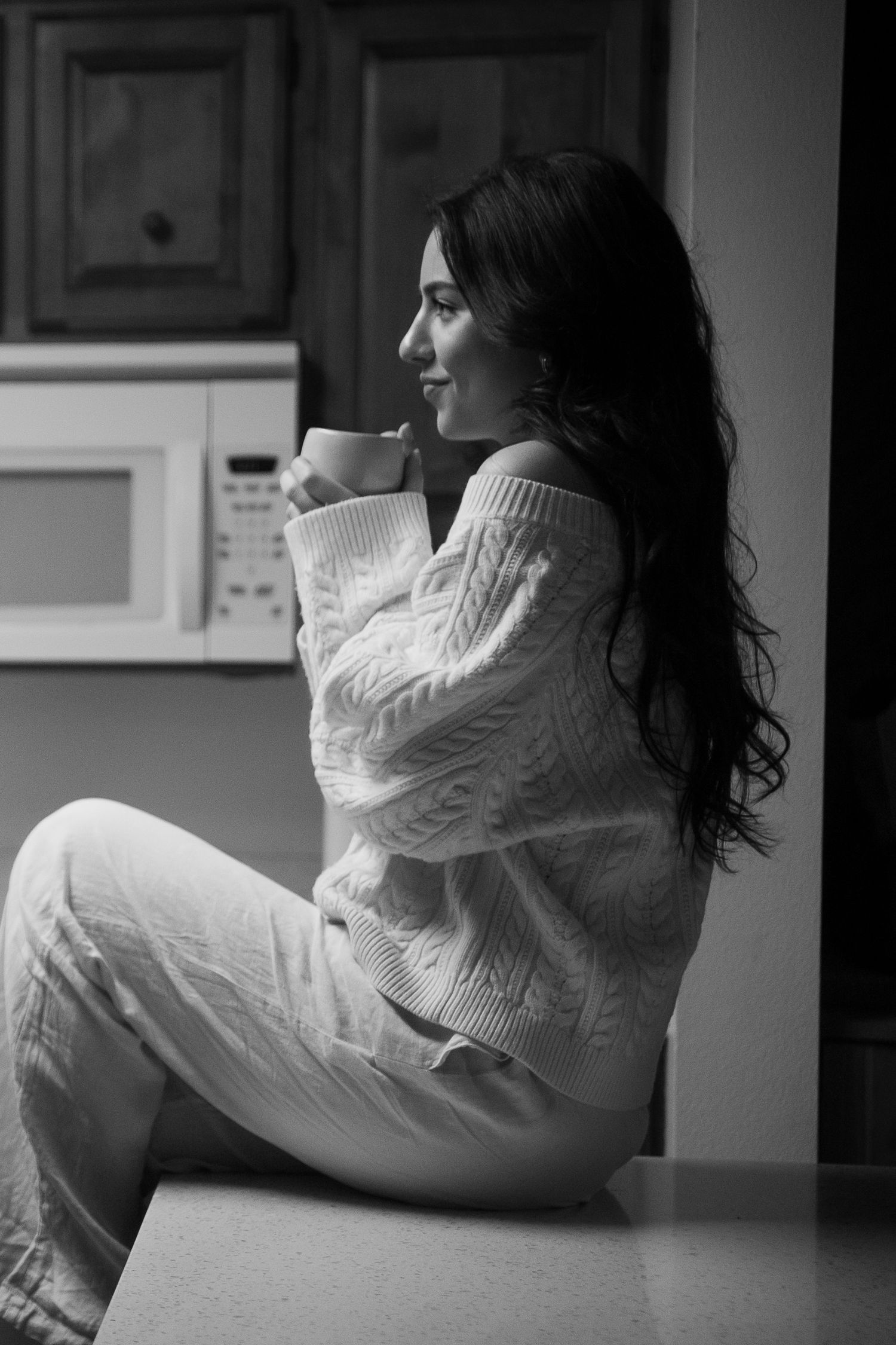 Black and white photo of a woman in a cozy knit sweater sitting on a counter, holding a mug in a kitchen.