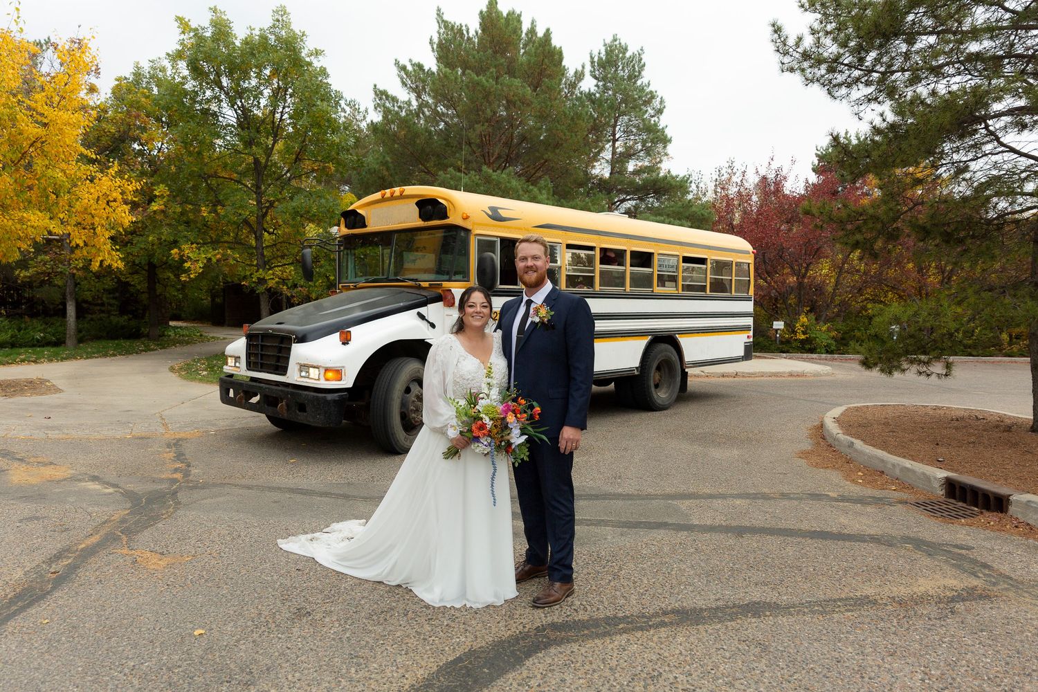 Bride and groom posing in front of a vintage yellow school bus during fall outdoor wedding celebration.