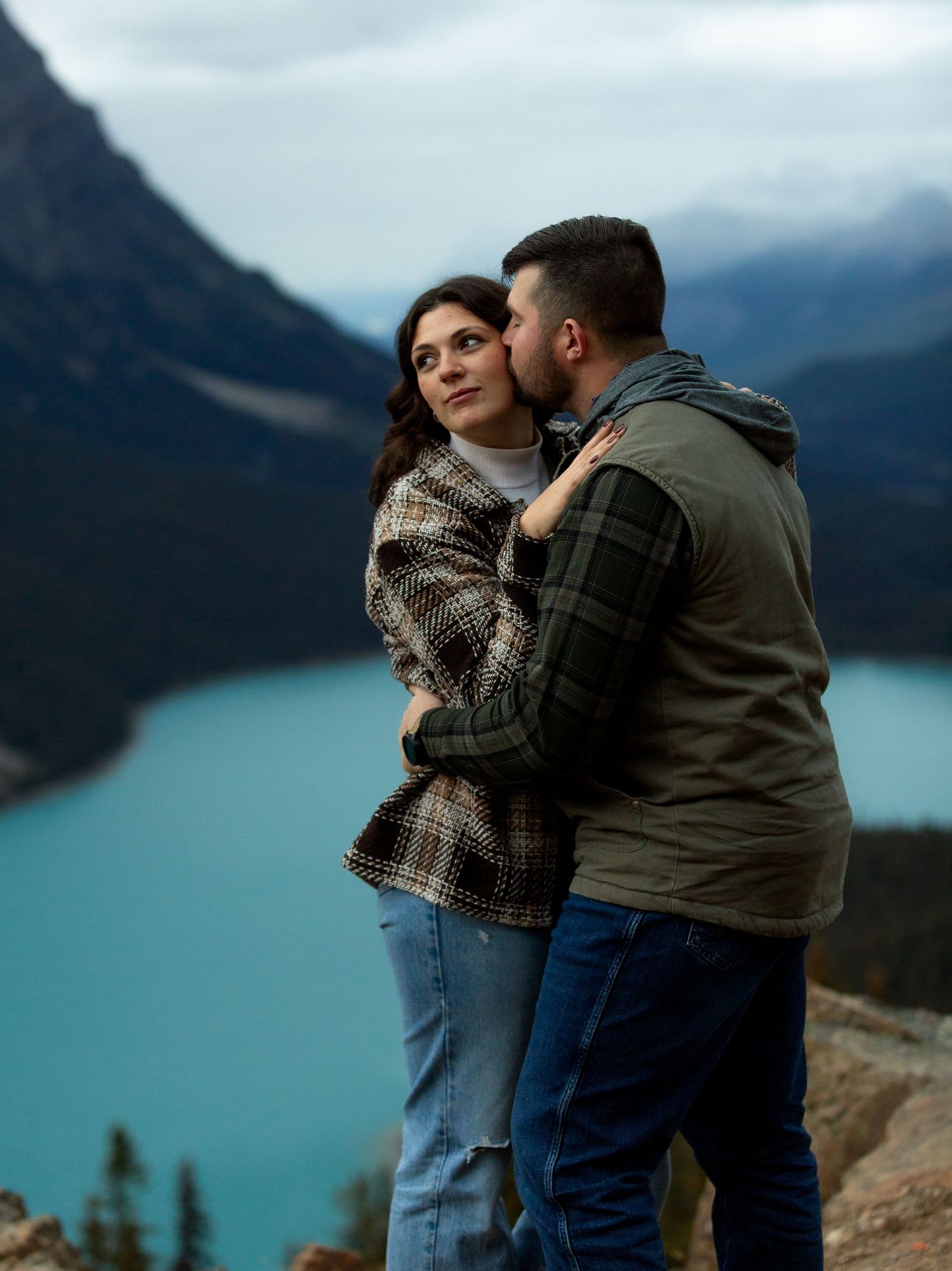 Couple embracing at Peyto Lake viewpoint in Banff, man kissing woman's cheek with turquoise lake and mountains behind.