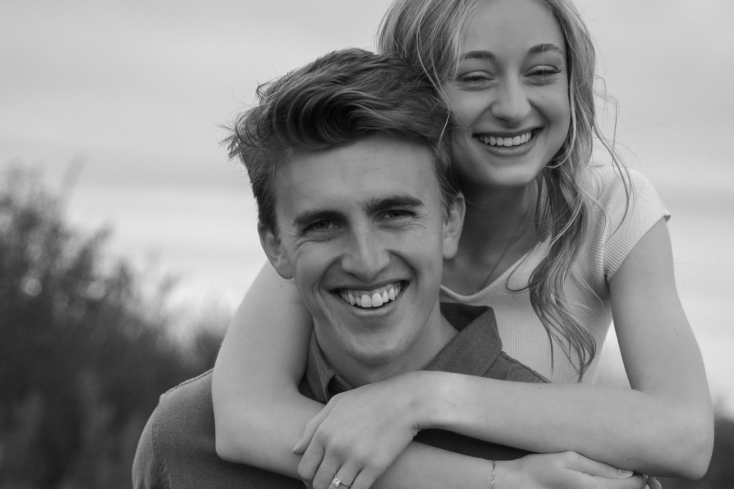 Black and white photo of a smiling young couple, woman hugging man from behind outdoors.