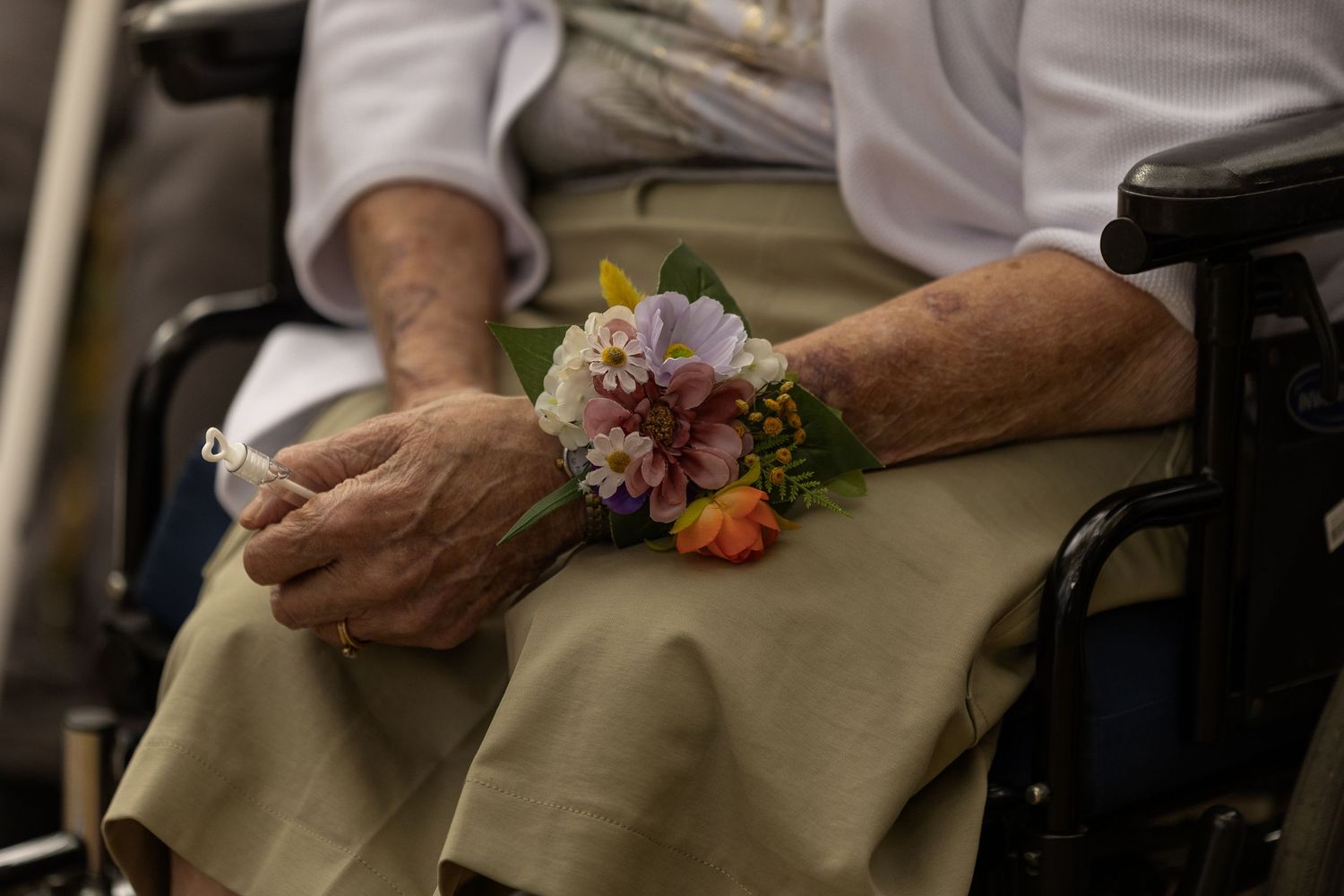 Elderly person in wheelchair with colorful floral wrist corsage resting hands on lap in beige skirt.