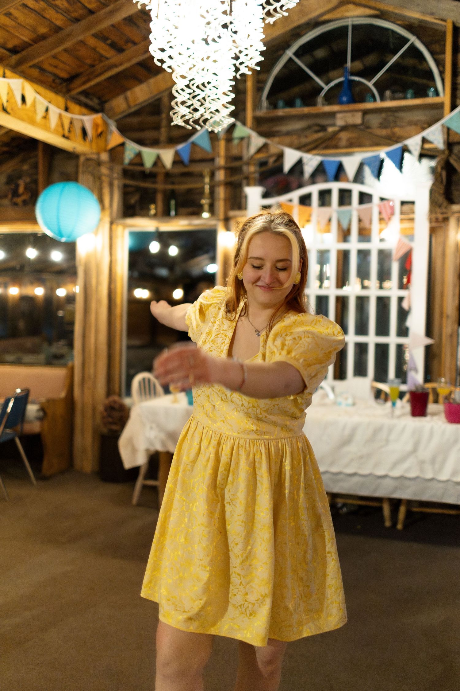 Young woman in yellow floral dress dancing joyfully at a rustic decorated party venue with string lights and bunting.