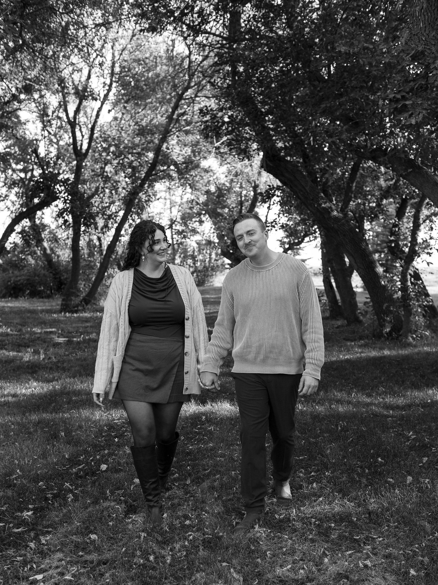 Couple holds hands while walking through wooded park in black and white.