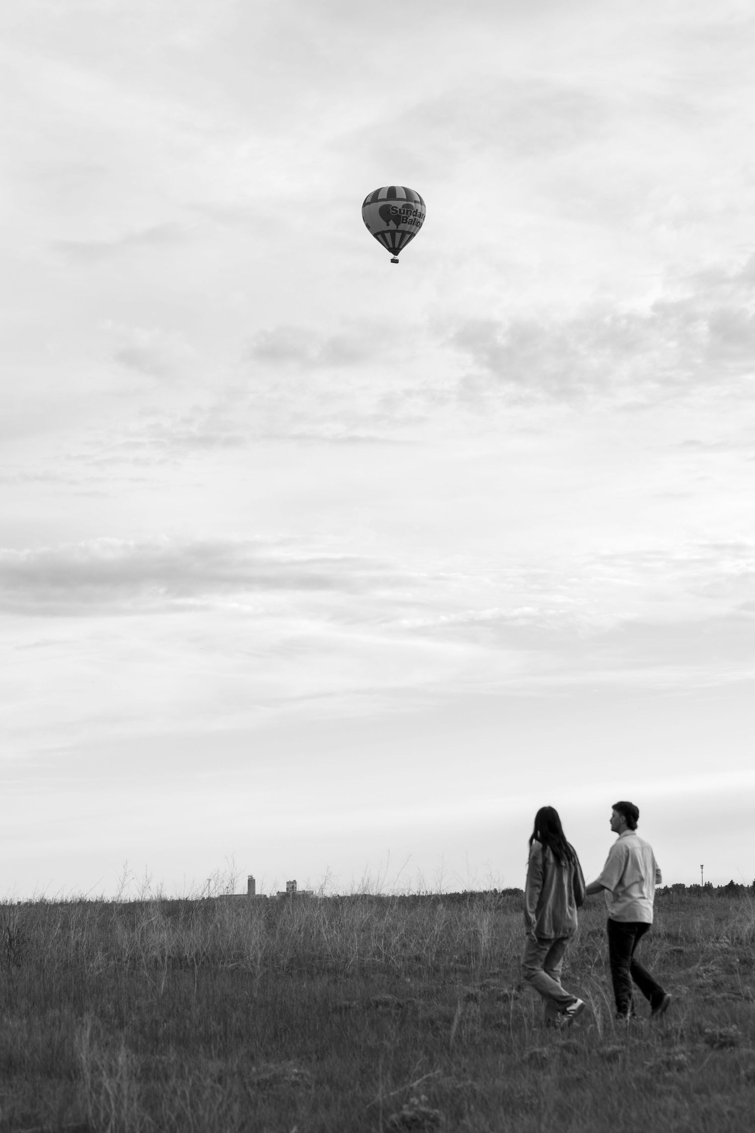Black and white photo of couple walking in field watching hot air balloon soaring in overcast sky.